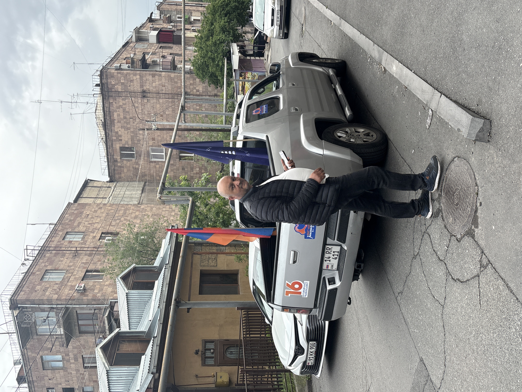 The image shows a man standing on a street next to two parked cars, likely in Yerevan, Armenia. The man is bald, wearing a black jacket, and holding a mobile phone in his left hand. Both cars are decorated with Armenian flags and political stickers, including the number "16" and the phrase "ՔԱՂԱՔԱՑԻԱԿԱՆ ՊԱՅՄԱՆԱԳԻՐ" (Civil Contract). The license plates read "91 KK 900" and "14 RR 411." The street is an urban environment with buildings in the background. The pavement is cracked and there is a manhole cover near the man's feet.