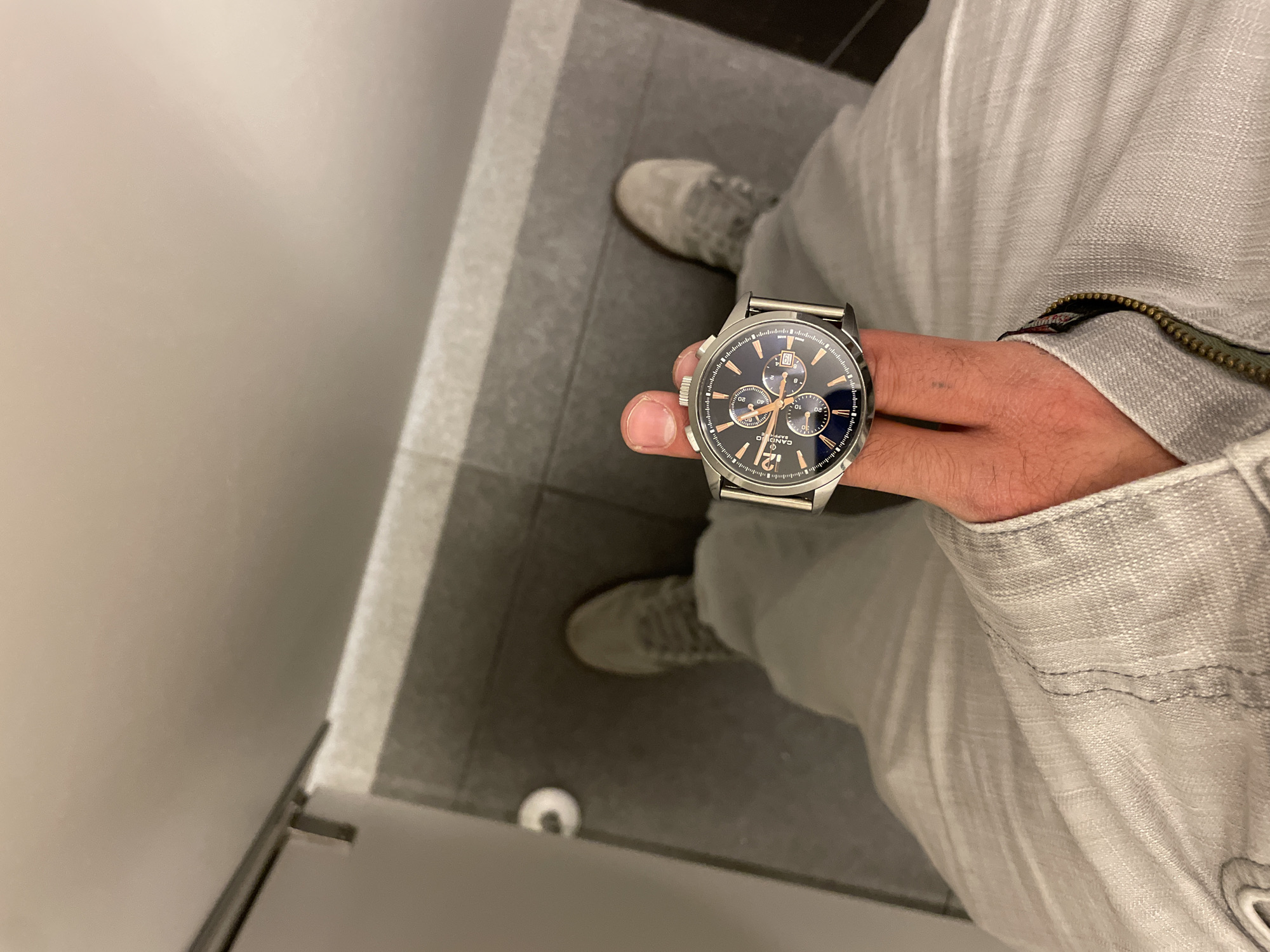 The image shows a person from a top-down perspective in an indoor setting. The person is wearing light gray pants, a light gray jacket, and gray suede shoes. The person is holding a silver wristwatch with a blue face. The background consists of gray tile flooring and a white wall. A toilet paper holder is visible on the floor. The location is a bathroom. The location in the city is Unknown, Unknown.