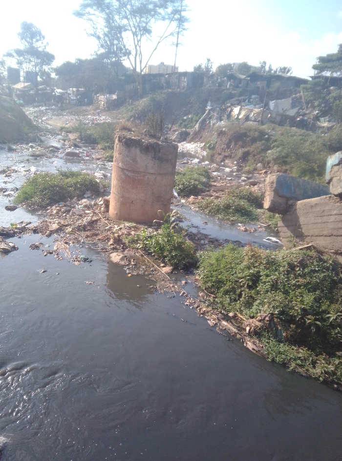 The image displays a highly polluted waterway, identified as a river or large stream, flowing through an informal settlement in Nairobi, Kenya. The water is dark grey, indicating heavy contamination, with a visible accumulation of floating debris including plastic bags, bottles, and general refuse. A prominent, broken concrete cylindrical pillar stands partially submerged in the water in the mid-ground, surrounded by clustered trash and green vegetation. On the right bank, closer to the foreground, multiple large, rectangular concrete blocks are visible, also interspersed with debris. Both banks of the waterway rise into a landscape densely populated by informal housing structures, primarily constructed from corrugated metal and other salvaged materials. These settlements extend into the hazy background under a bright sky, with several trees visible on the distant horizon. No individuals or specific human actions are discernible within the frame. The scene presents evidence of significant urban waste accumulation and environmental degradation.