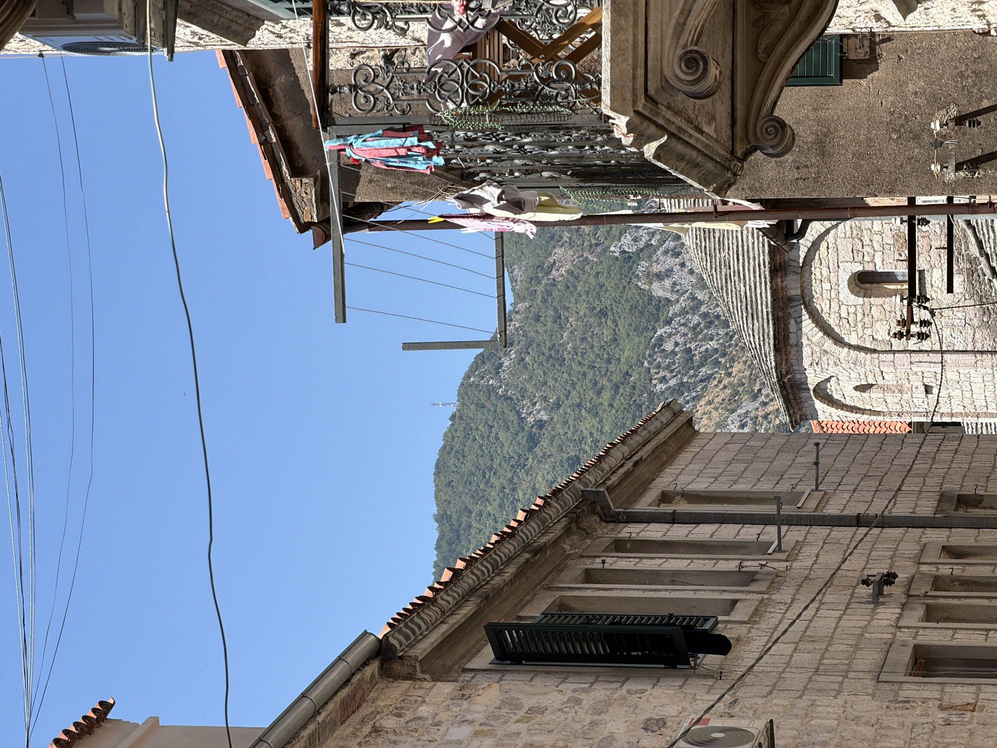 This image depicts a street scene in Kotor, Montenegro. The foreground shows the exterior of two buildings constructed with light-colored stone. The building on the left has a tiled roof and a partially open louvered window. An air conditioning unit is mounted to the exterior wall. The building on the right features a detailed balcony with ornate ironwork. Laundry is hanging on a drying rack attached to this building. In the mid-ground, a stone building with a domed roof and arched windows is visible. A clothesline stretches across the alleyway. The background is dominated by a steep, tree-covered mountain. The sky is clear and blue, with some electrical wires strung across the upper portion of the image. The overall impression is of a sun-drenched, historic European town.
