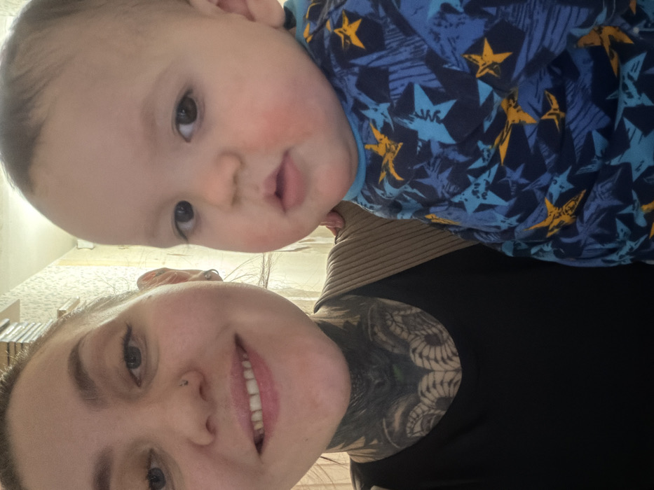 A person with light skin and a neck tattoo smiles at the camera. A baby, with chubby cheeks and brown eyes, is held above the person. The baby wears a blue patterned shirt with yellow stars. The adult wears a black top over a ribbed beige undershirt. The photo is taken at an angle, with the subjects appearing sideways.