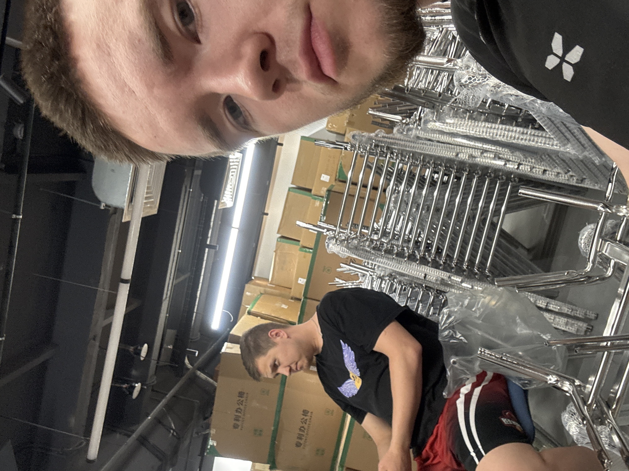 The photo appears to have been taken indoors, possibly a warehouse or storage area, in the city of Kazan, Russia. The photo contains two men, one is looking down and another is taking a selfie. The man taking the selfie is at the top left of the image, and wearing a black shirt with a white logo. The other man is at the bottom center of the image and is wearing a black T-shirt with a purple design and red shorts with white stripes. Behind the men are stacks of metal chairs, packed in plastic. Behind the men are multiple cardboard boxes with Chinese writing on them. There is a fluorescent light on the ceiling.