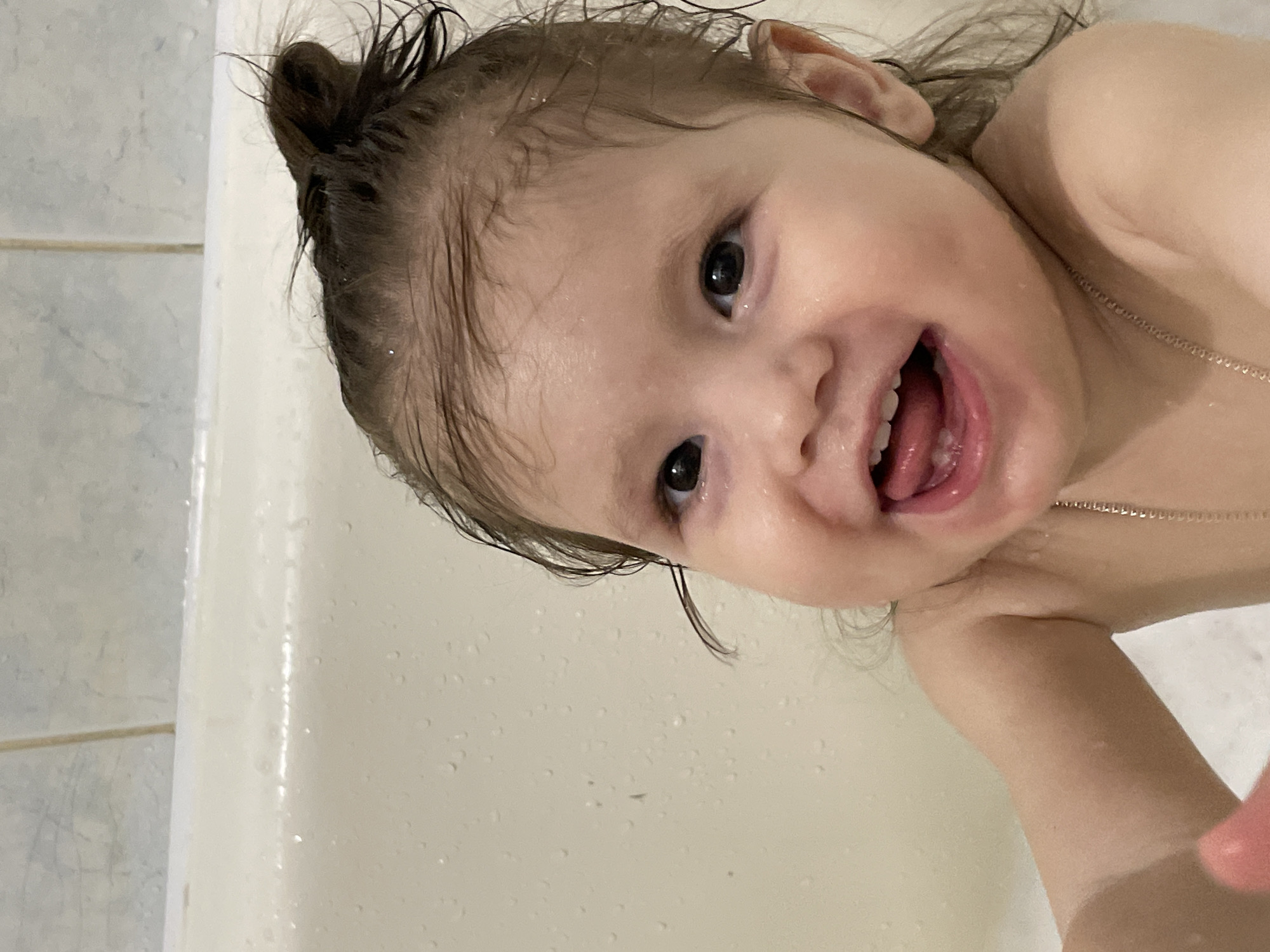 A close-up shot reveals a child in a bathtub. The child is fair-skinned with wet, dark hair pulled into a small bun on top of their head. Water droplets are visible on the child's face and hair. The child is smiling widely, revealing their teeth and tongue. A delicate necklace is visible around the child's neck. The bathtub is white, and the background includes light-colored tiles. There is no indication of the image being taken in Izhevsk, Russia.