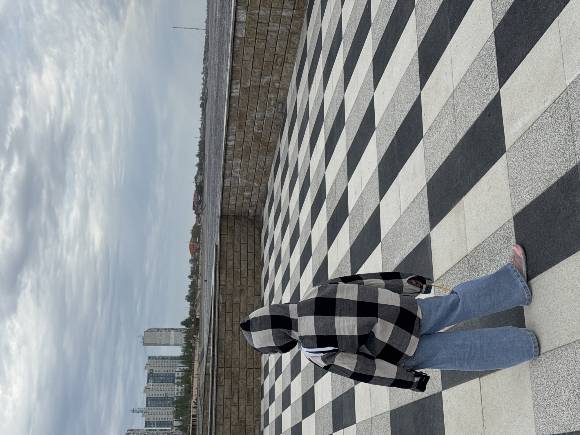 A person wearing a black and white checkered hooded jacket and blue jeans is standing on a black and white checkered tiled surface. The scene is set with a gray, cloudy sky and buildings in the distance. A low brick wall runs along the left side of the frame. The location is most likely in Nizhnevartovsk, Russia, inferred from the architectural style of the buildings.