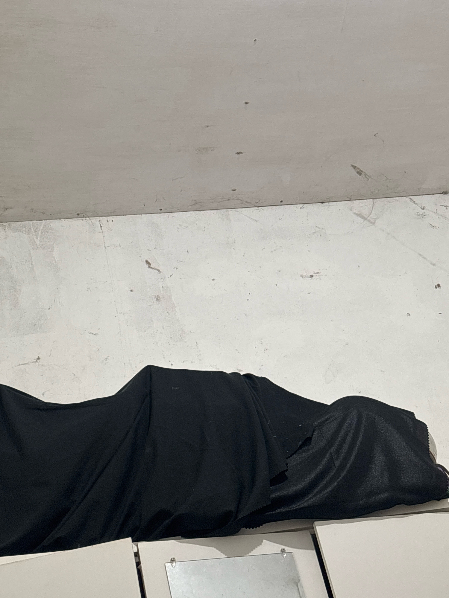The image depicts a room with white, textured walls and a ceiling. A black fabric is draped over a form, obscuring its details. The fabric is wrinkled and folded, suggesting it is covering an object or possibly a person. The floor area visible in the lower portion of the image appears to be white and flat, possibly a tabletop or shelves, with a small metal object attached to one of them. The overall lighting is dim and appears to be artificial. The scene is nondescript and could be any indoor space. There is no identifiable context that places it specifically in North Jakarta, Indonesia, or any other urban location.