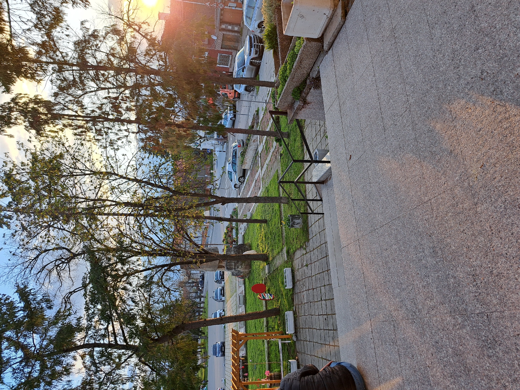 The image depicts an outdoor scene in Lokorsko, Bulgaria, during sunset. The foreground shows a section of a granite-tiled elevated platform or patio. To the left, a grassy area with several mature trees stretches towards the street, which is lined with parked cars. Visible in the grassy area are a wooden structure that may be a play area, a red and white sign, and several rectangular concrete planters. The setting sun casts a warm glow on the scene, particularly on the granite surface and the upper floors of buildings across the street. The sky is a mix of blue and clouds. A partial view of a person's jacket and jeans can be seen at the bottom edge of the image.