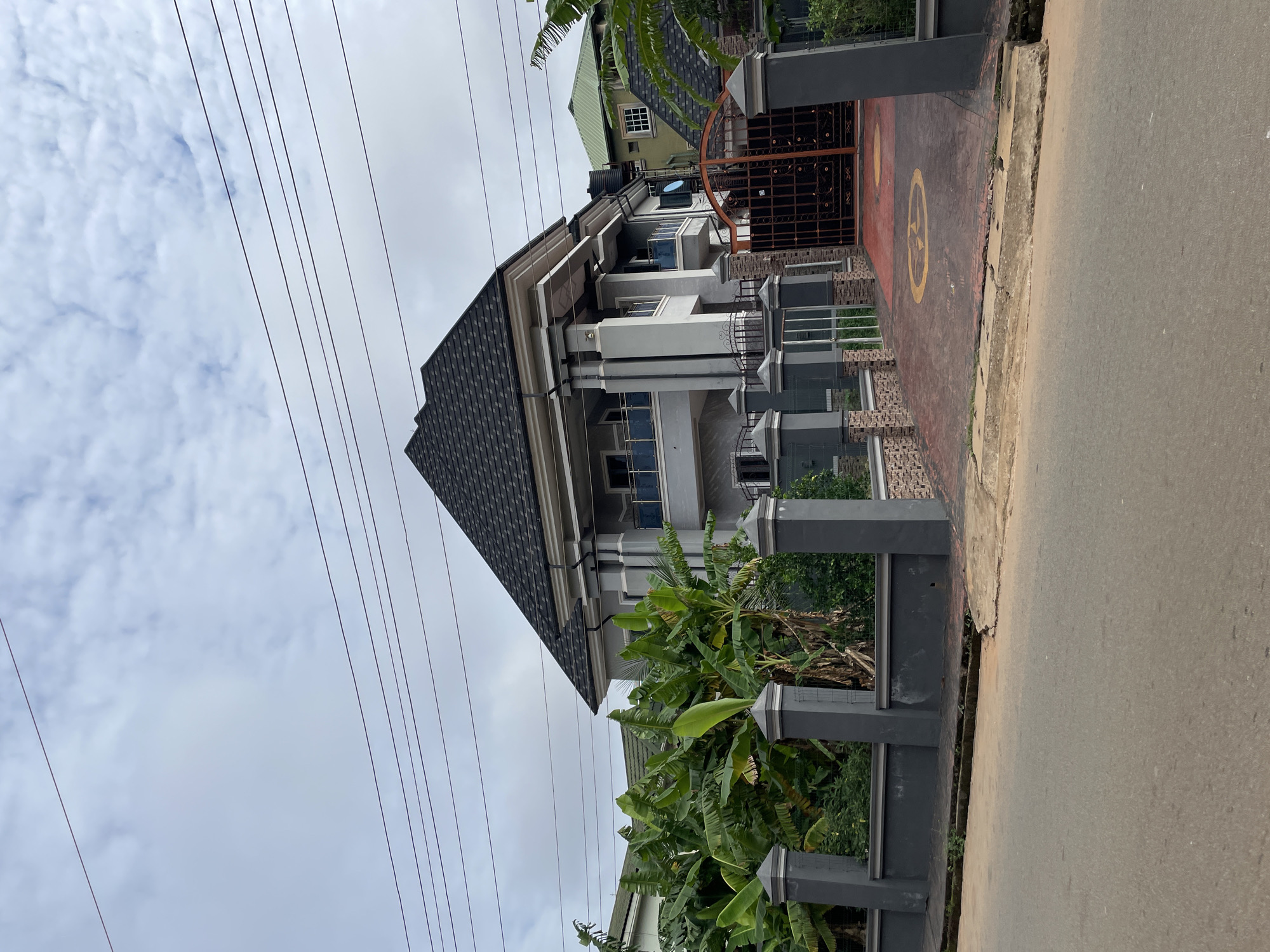 The image captures a modern multi-story residential building in Onitsha, Nigeria. The house has a dark gray roof and white walls with gray accents. An elaborate gray metal gate secures the property. A gray fence borders the property along the street, with dense green foliage growing within. Power lines stretch across the sky above. The scene is captured during daylight hours under a cloudy sky. The street in front of the house is paved.
