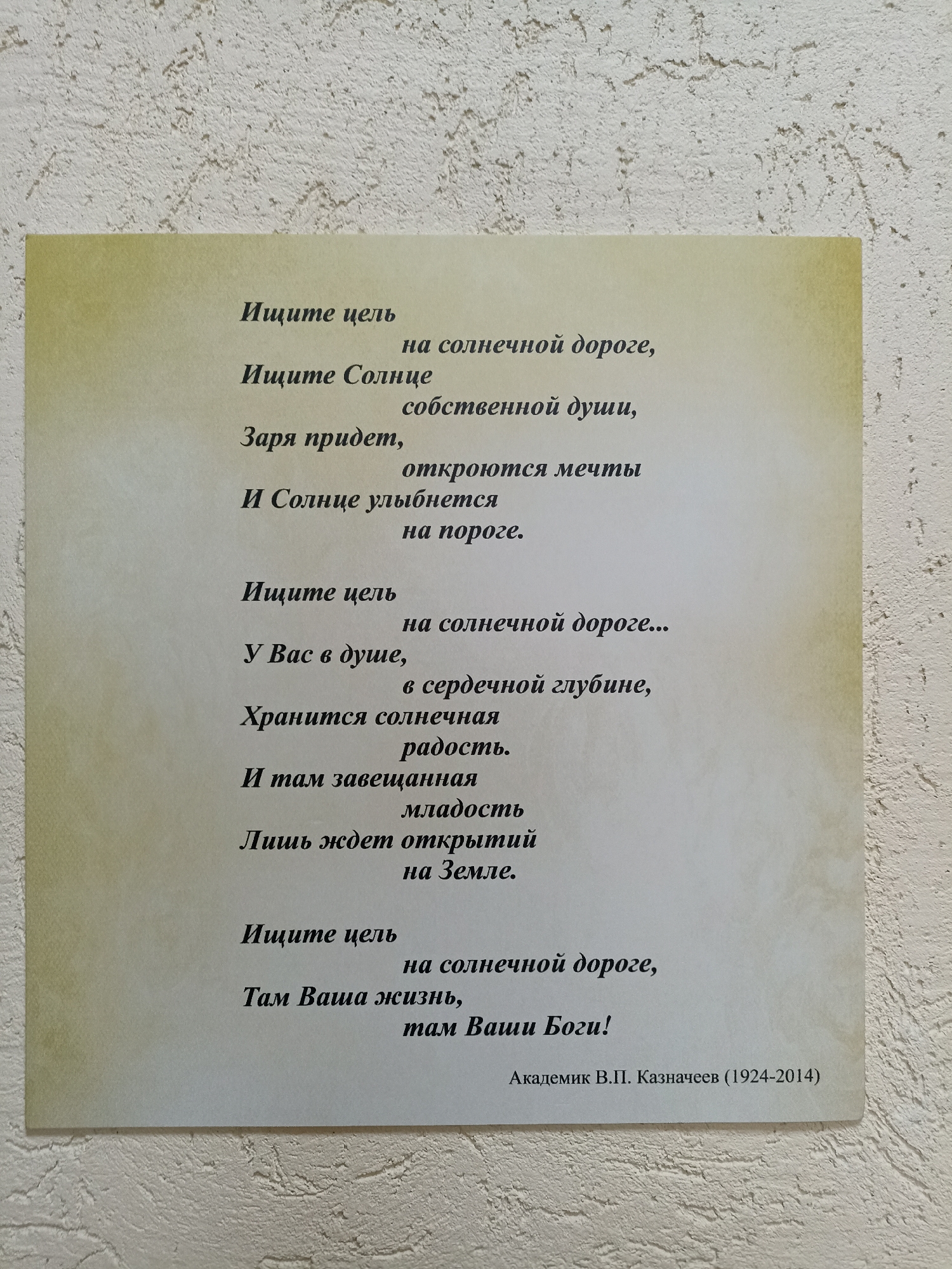 The image shows a poem printed on a light-colored card, which is mounted on a textured wall. The poem is in Russian and appears to be motivational in nature. The text is dark and is arranged in several stanzas. At the bottom of the card, there is a name and dates, "Академик В.П. Казначеев (1924-2014)", indicating the author. The background wall has a rough, off-white texture, typical of many interior walls. There are no people or specific locations within the image that would allow for identification of a city or even a specific type of building. The scene is an indoor setting, likely a home or an office, where this printed poem is displayed.