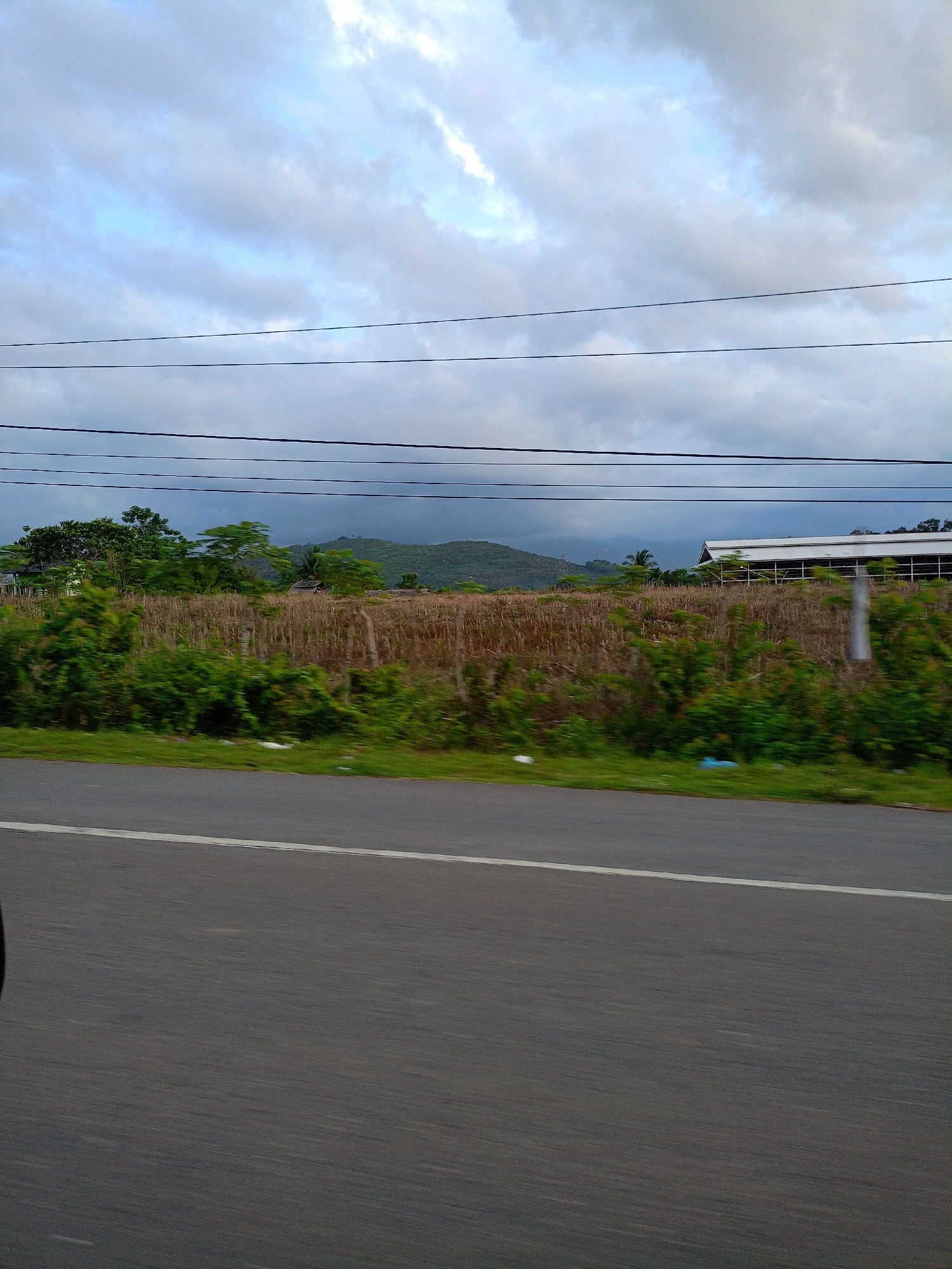 The photograph captures a roadside scene near Lhokdagang, Indonesia under a cloudy sky. The image, likely taken from a moving vehicle, features a two-lane road marked with a white line. Adjacent to the road, a grassy verge transitions into a dense, overgrown area with bushes and tall, dried vegetation. In the background, a long, low structure with a metallic roof is visible, partially obscured by the vegetation. Beyond this, a gentle, forested hill rises in the distance. Overhead, several utility lines stretch across the sky. The image conveys a sense of motion and the rural landscape surrounding Lhokdagang.