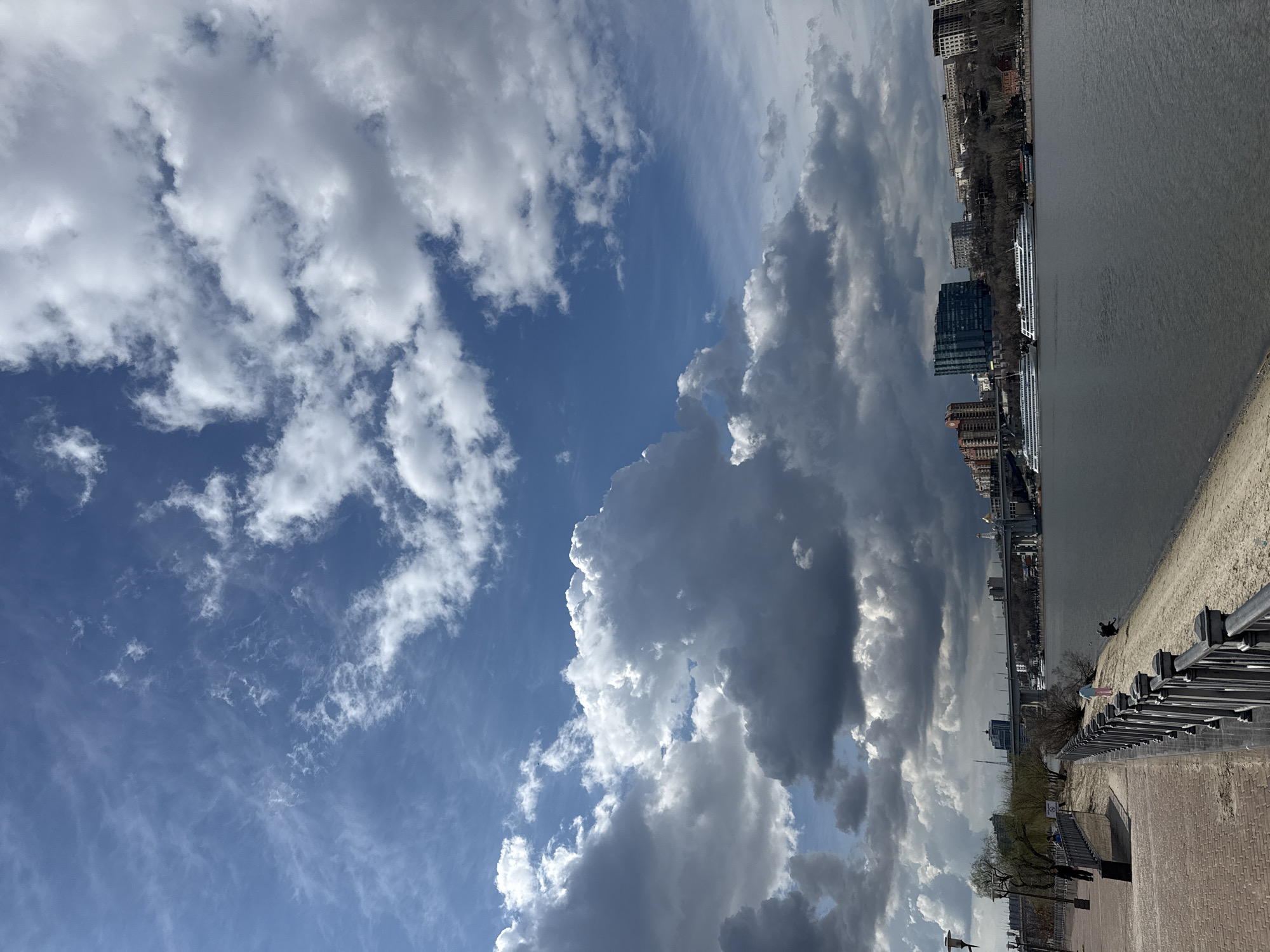 The image depicts a daytime view of Rostov-on-Don, Russia, from a riverside perspective. The sky occupies a significant portion of the frame, featuring a mix of blue sky and cumulus clouds, some appearing white and fluffy while others are darker and more ominous. The city skyline is visible along the right side of the image, showcasing buildings of various heights and architectural styles. A body of water, likely the Don River, stretches along the right edge, with a sandy or concrete embankment visible in the foreground. A metal railing runs along the embankment. A few people are discernable along the embankment, appearing as small figures in the distance. Overall, the image presents a scene of contrasting elements – the dynamism of the sky juxtaposed with the static cityscape.