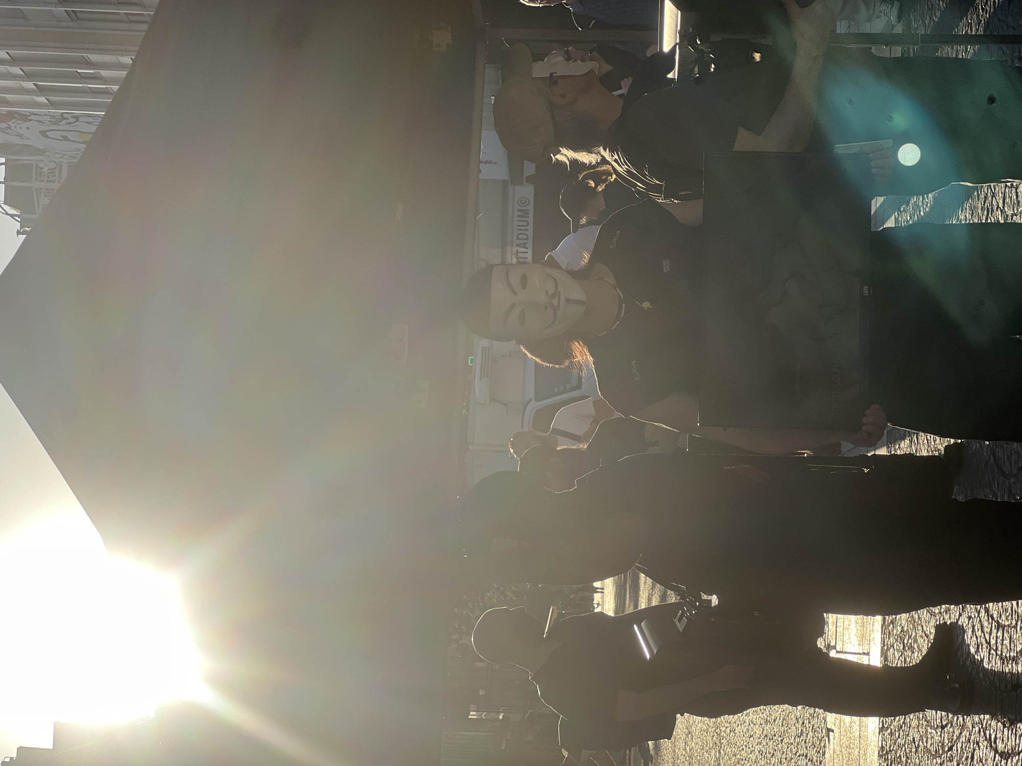 The image depicts a group of people standing on a cobblestone street, one of whom is wearing a Guy Fawkes mask. The word "STADIUM" is visible on the side of a vehicle in the background. The location is likely in Paris, France, based on the architectural style and cobblestone streets. The photo is taken against the sun, creating a significant glare that obscures details.