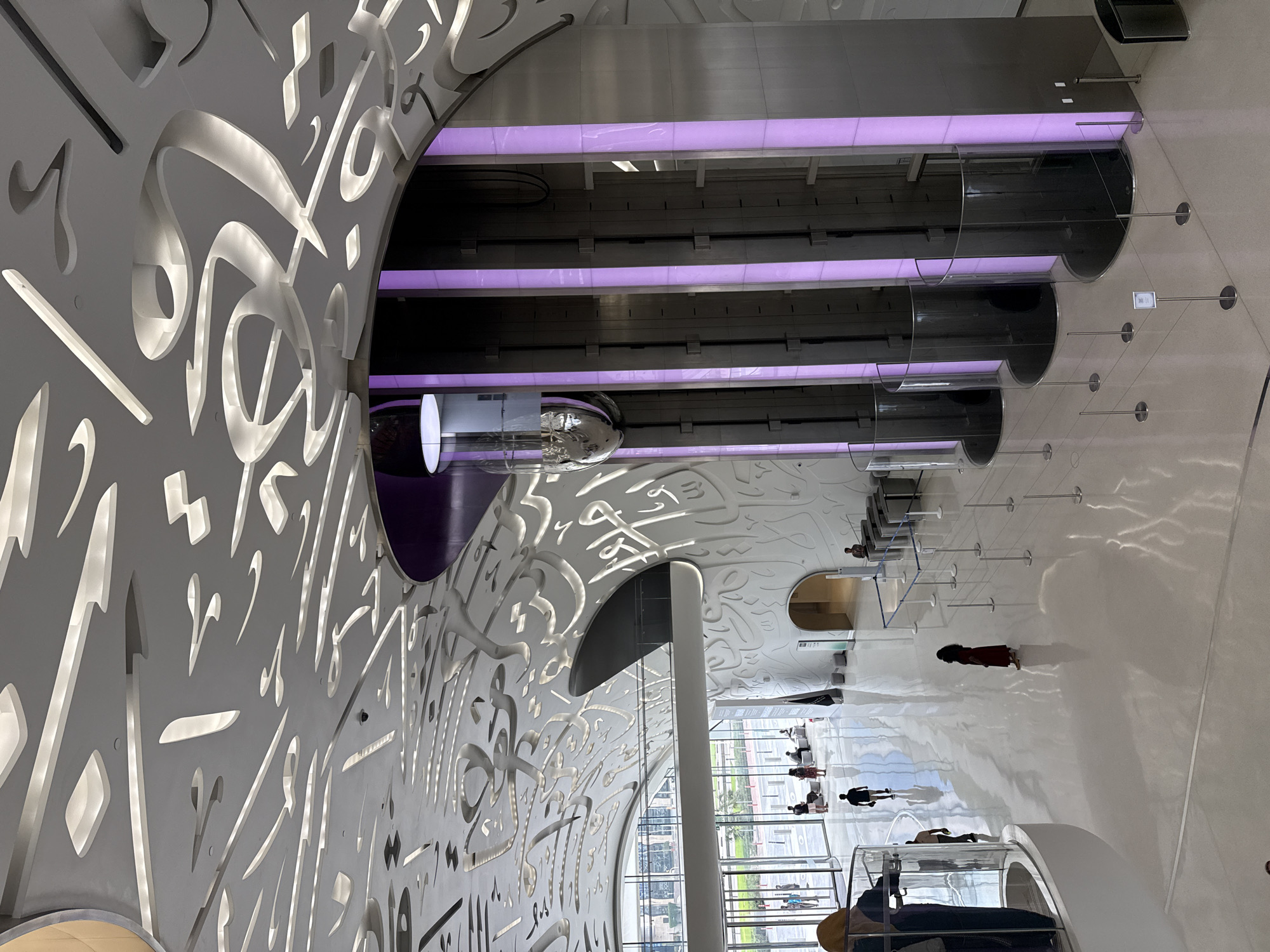 The image depicts the interior of the Museum of the Future in Dubai, United Arab Emirates. The distinctive curved walls are adorned with Arabic calligraphy cutouts, illuminated from within. A series of cylindrical, glass-enclosed elevators run vertically, lit by horizontal purple bars. People are walking on the shiny white floor. The scene appears to be a view looking down towards the ground floor through an opening in the structure. A large pillar is visible along with windows looking out to the exterior of the building. There are people interacting with displays along the windows.