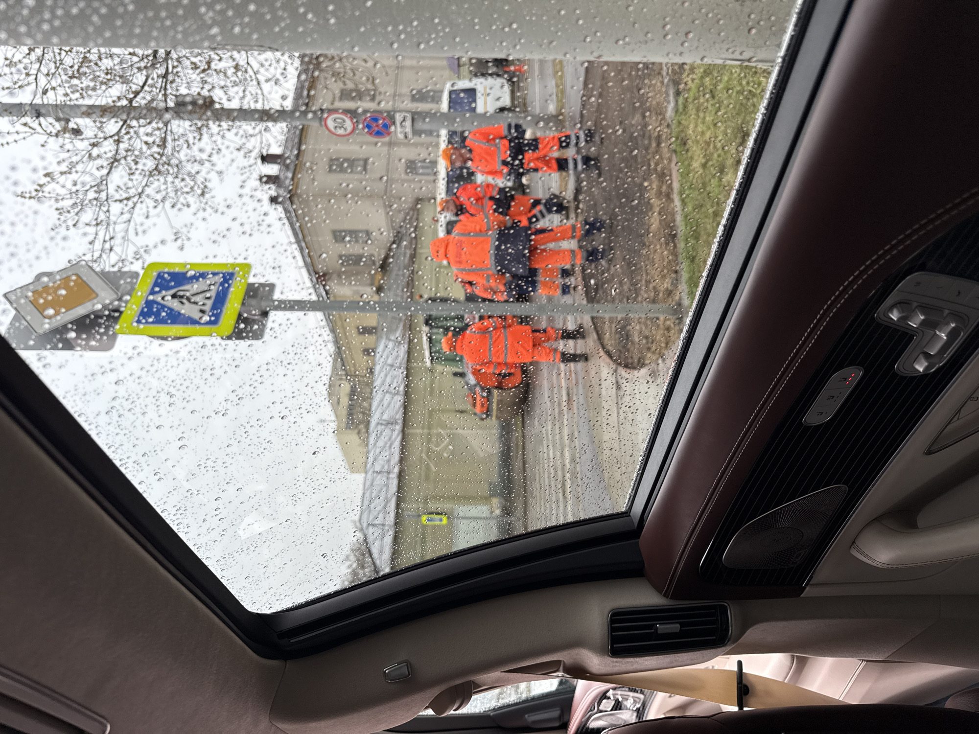 The image, taken from inside a car through an open sunroof on a rainy day, shows a street scene in Moscow, Russia. Several road signs are visible. A group of workers wearing bright orange jackets and dark pants are working on the street. A white van is parked nearby. The car's interior includes brown leather trim, and air vents.