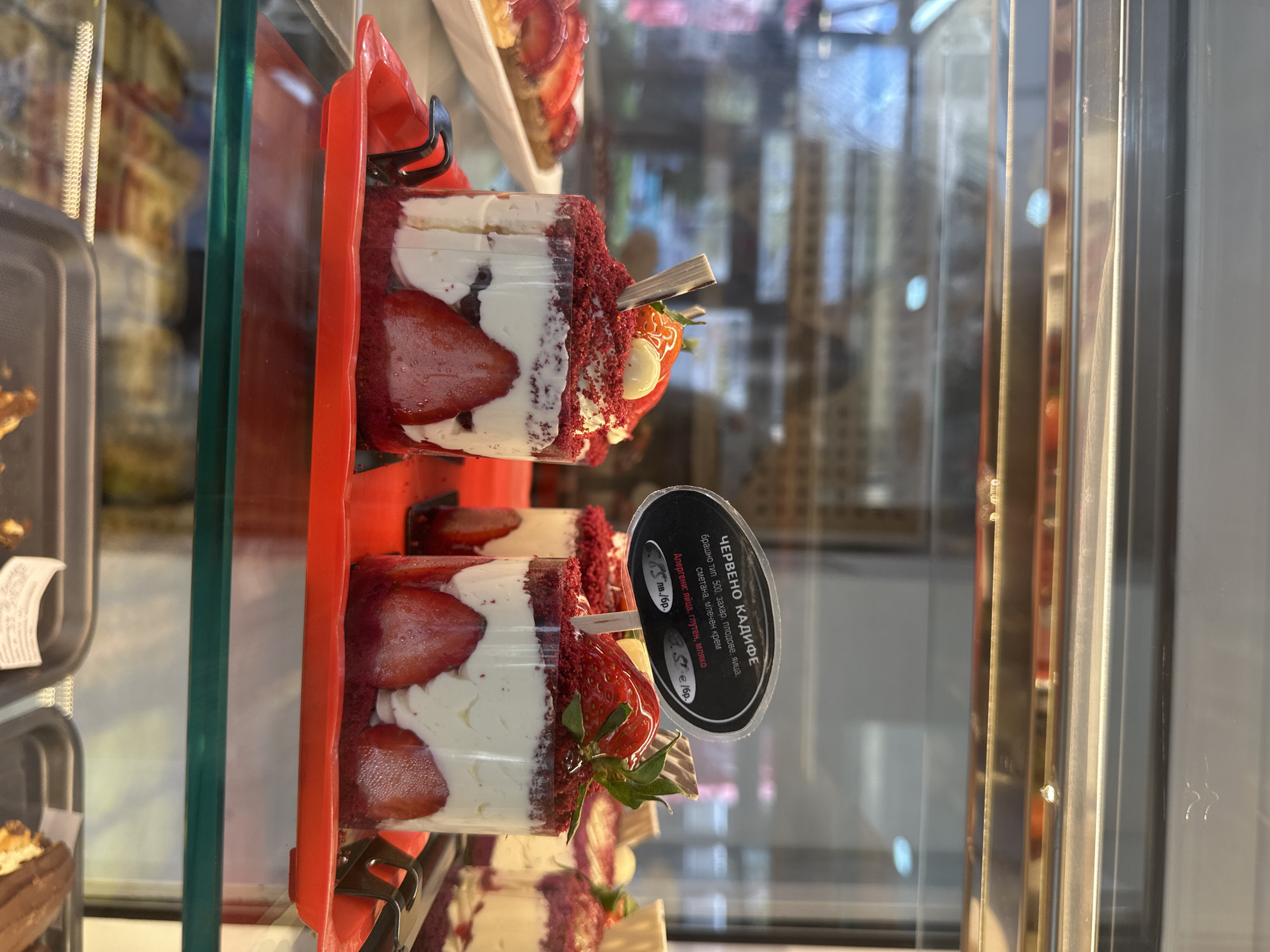 A vertically oriented, close-up photograph captures two individual "Red Velvet" desserts displayed in a refrigerated glass case, likely within a bakery or cafe in Sofia, Bulgaria. Each dessert is presented in a clear plastic cup, showcasing distinct layers of vibrant red cake, fluffy white cream, and fresh sliced strawberries. The top dessert is further garnished with red cake crumbs and a whole strawberry, which is drizzled with white chocolate and skewered by a small wooden pick.
Attached to the upper dessert is a black, oval-shaped tag providing information in Bulgarian. The text clearly identifies the dessert as "ЧЕРВЕНО КАДИФЕ" (Red Velvet). Ingredients are listed as "брашно тип 500, захар, плодове, яйца, сметана, млечен крем" (flour type 500, sugar, fruits, eggs, sour cream, milk cream). Allergens are also specified: "яйца, глутен, мляко" (eggs, gluten, milk). The price is visible as "6 лв./бр." (6 Bulgarian Leva per piece) and "3.50 €/бр." (3.50 Euros per piece).
The desserts rest on bright red plastic trays inside the display unit. The glass enclosure features prominent reflections of the surrounding indoor environment and metallic vertical supports. Other out-of-focus items, possibly more desserts or trays, are partially visible in the background and to the left. The scene evokes the typical presentation of tempting sweets available for purchase.