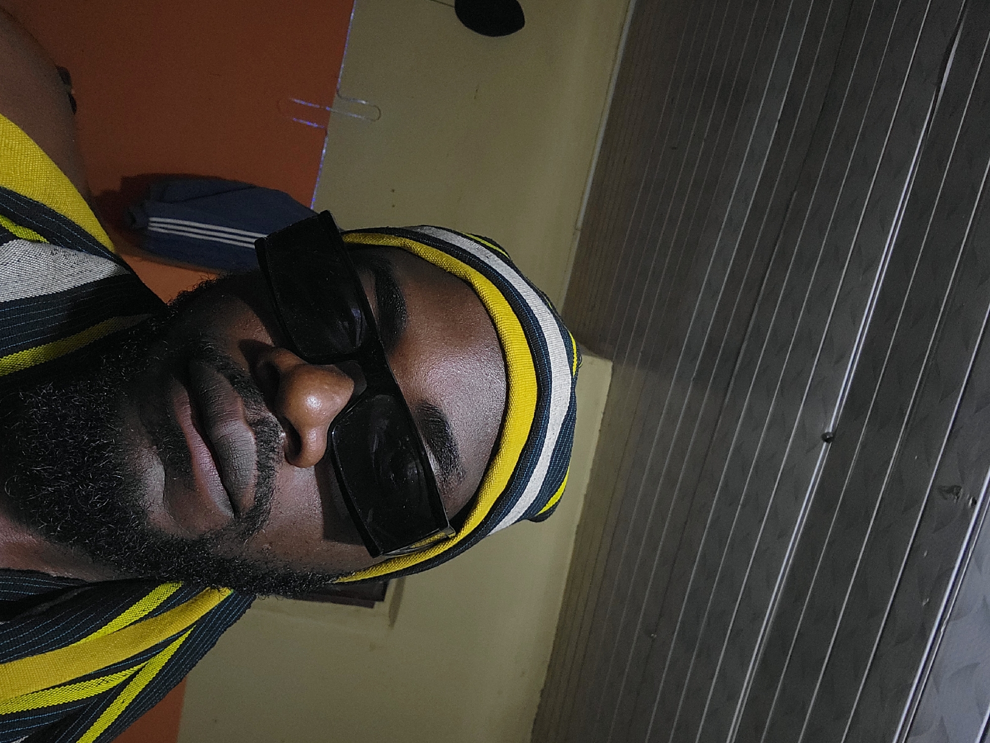 A man is looking at the camera, wearing sunglasses, a beanie and a scarf. The beanie and scarf are yellow, black and white striped. The man has a beard and a mustache. He is wearing a black t-shirt. The background is a room with a wooden ceiling, orange walls and a blue and white striped jacket hanging on the wall. There is also a black microphone stand on the left side of the image. The image was taken in Jos, Nigeria.