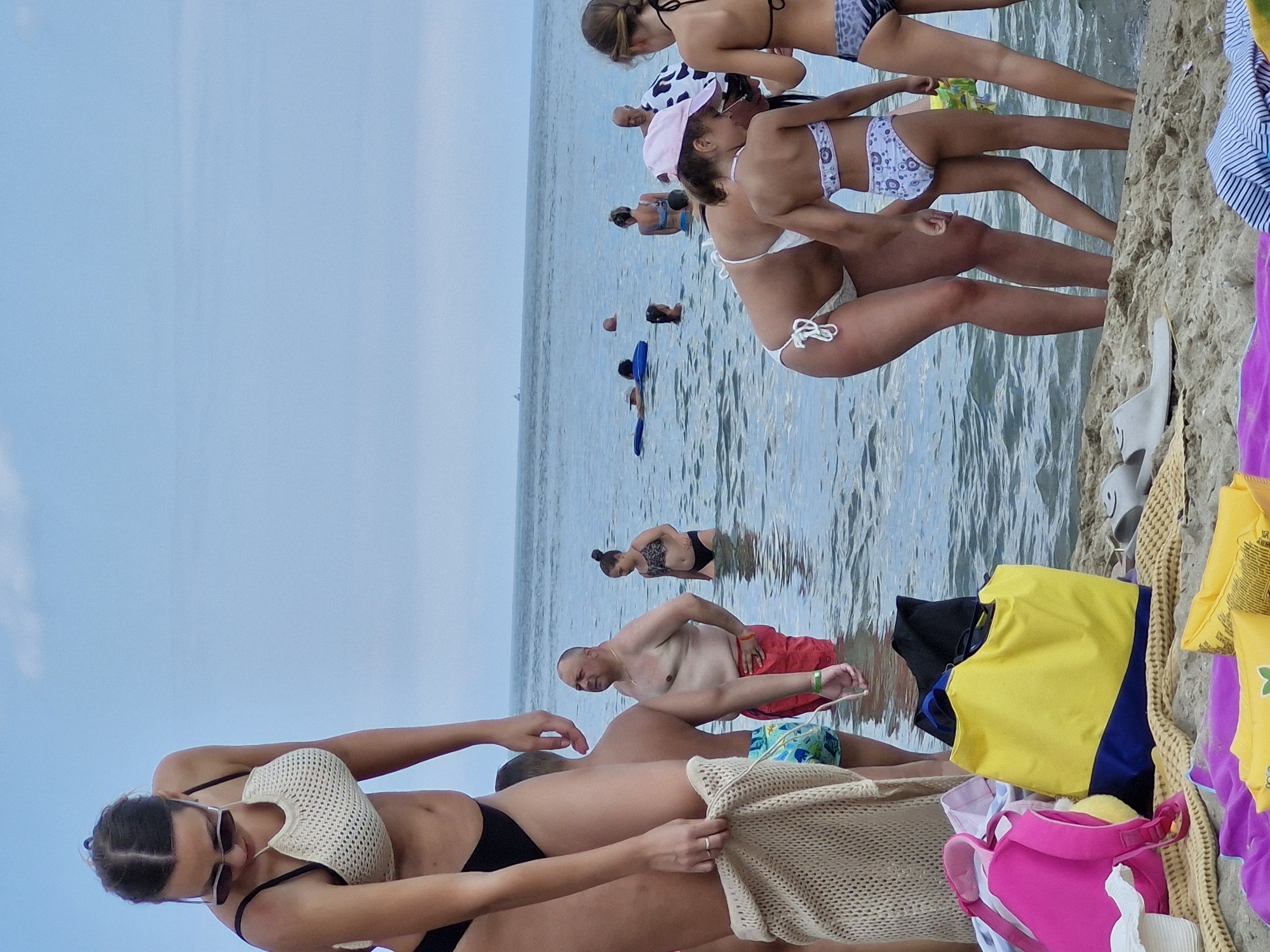 This media file shows a lively scene at Sveti Vlas, Bulgaria, likely on a bright and somewhat overcast day at the beach. The water is a calm, clear blue, reflecting the sky. The foreground is dominated by the sandy shore and a pile of colorful beach items, including a bright yellow vest, a pink bag, and other assorted clothing.
In the water, several people are enjoying the day. A man in red shorts is lying down, and further out, other individuals are swimming or wading. The middle ground features a group of people, primarily women and children, in swimwear. One woman is lying on the sand, while others are either standing or in the shallow water. Two young girls are particularly prominent, one in a white bikini with a patterned trim and the other in a similar white bikini. Another woman is wearing a patterned bikini.
In the background, more people are visible in the water, suggesting a moderately busy beach environment. The overall atmosphere appears relaxed and recreational, with people engaged in typical beach activities like swimming and lounging. The presence of beach towels, clothing, and personal items indicates a day spent relaxing and enjoying the seaside. The lighting suggests it might be late morning or early afternoon, with the sun partially obscured by clouds.