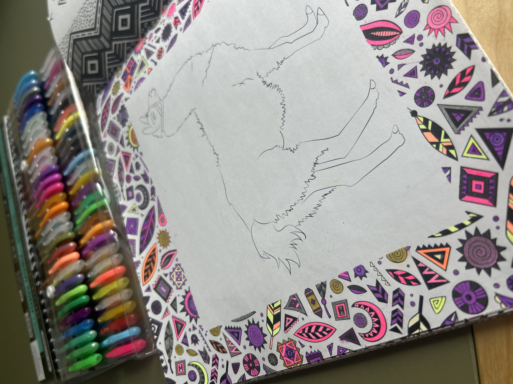 This close-up shot captures an open coloring book and a set of gel pens, suggesting a creative activity. The coloring book is open to a page featuring a large, uncolored outline drawing of an animal, likely a llama or an alpaca, positioned centrally. The animal is depicted with a stylized head, small ears, and a textured body, possibly wearing a small decorative element on its head.
Surrounding the central animal outline, the page's border is filled with intricate, abstract geometric and organic patterns. Many of these patterns are already colored in vibrant, contrasting hues, including neon pink, bright yellow-green, purple, orange, and black, alongside some grey and golden tones. The designs feature triangles, squares, circles, swirls, feather-like shapes, and sunburst motifs.
To the left of the coloring book, a clear plastic case holds numerous gel pens or markers, arranged neatly in rows. The pens display a wide array of colors, including various shades of blue, green, yellow, orange, pink, and purple, with translucent caps. Faint text "NEON COLOR" is visible on the side of the pen case, indicating the type of pens.
The scene is well-lit, possibly by natural daylight or bright artificial light. The image is taken from a slightly elevated perspective, looking down onto the items. The coloring activity appears to be in progress or recently undertaken, with the main animal figure awaiting coloration.