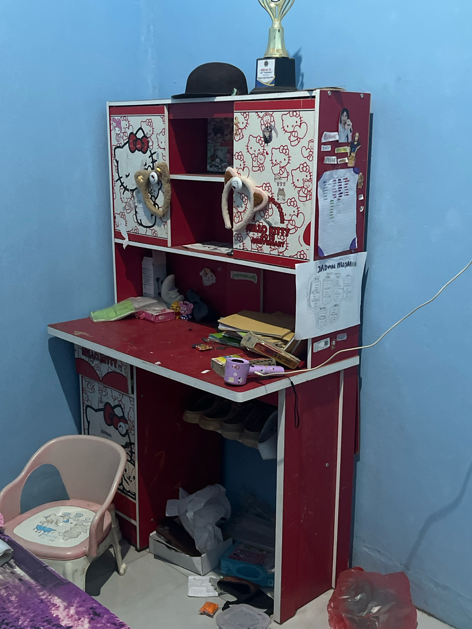 A red and white Hello Kitty themed desk with a hutch sits against a light blue wall in what appears to be a bedroom in Kebumen, Indonesia. The desk surface is cluttered with various items, including papers, pens, a purple cup, and a toy. The hutch contains shelves displaying more Hello Kitty decorations and a black bowler hat. To the left of the desk is a small pink chair. The floor is covered with a rug and some scattered items, including a red plastic bag and a white box. The overall scene suggests a child's or young person's personal space.