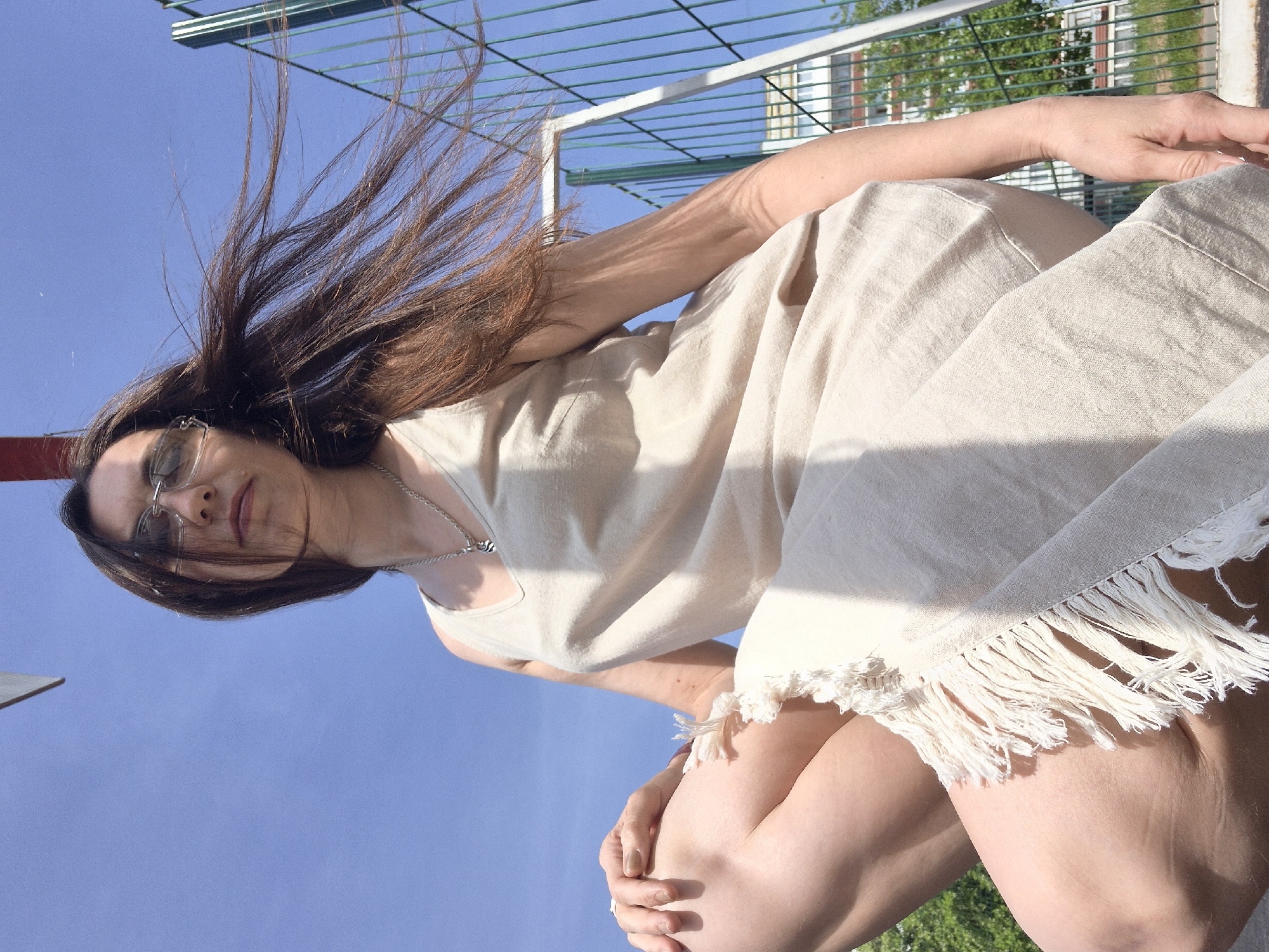 A woman is seen from a low angle looking downward. She has long brown hair, is wearing glasses and a silver necklace, and is dressed in a beige dress with fringed trim. She is outdoors on a sunny day with a blue sky. Behind her are green metal bars, part of a fence or enclosure, and some greenery, possibly trees. The location is an unidentified city, Unknown.