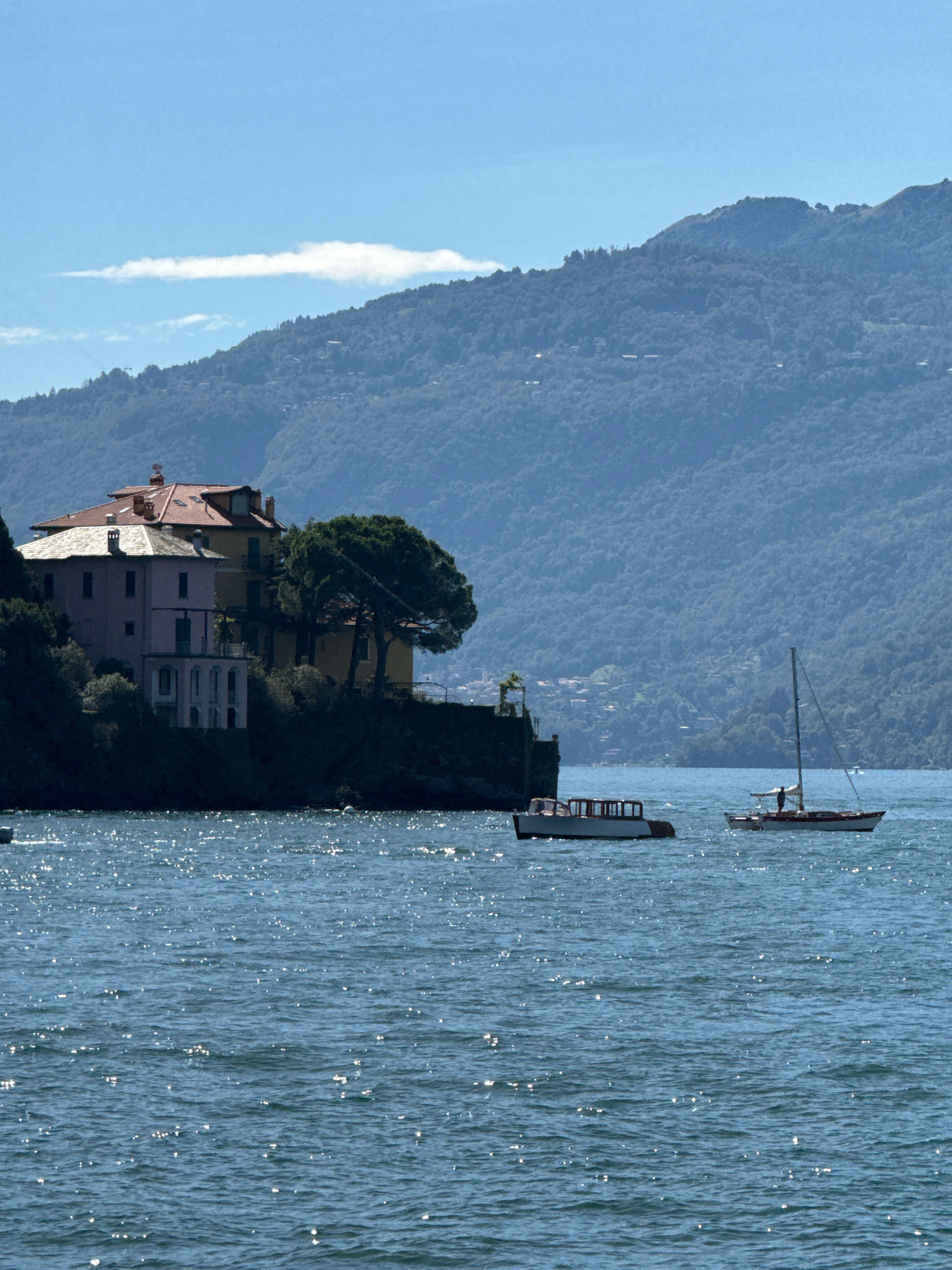 An image captured from a body of water shows a cluster of buildings situated on a rocky embankment. The buildings are multi-story structures with pitched roofs, one painted in shades of pink and yellow, with architectural details like balconies and windows. A large, mature tree with dark foliage stands beside the buildings.
In the foreground, the water is a deep blue, reflecting the sunlight with numerous bright sparkles. Two boats are visible on the water. The boat on the left is a white and brown vessel with a covered cabin area. The boat on the right is a sailboat with a tall mast, a single sail, and a person standing on its deck.
In the background, a lush green mountainside rises steeply, covered in dense foliage and dotted with scattered houses. A light blue sky with a wispy white cloud stretches above the mountain. The overall scene suggests a picturesque, possibly Mediterranean, location. The architectural style and the presence of a lake with surrounding mountains are characteristic of towns on Lake Como, such as Varenna, Italy.