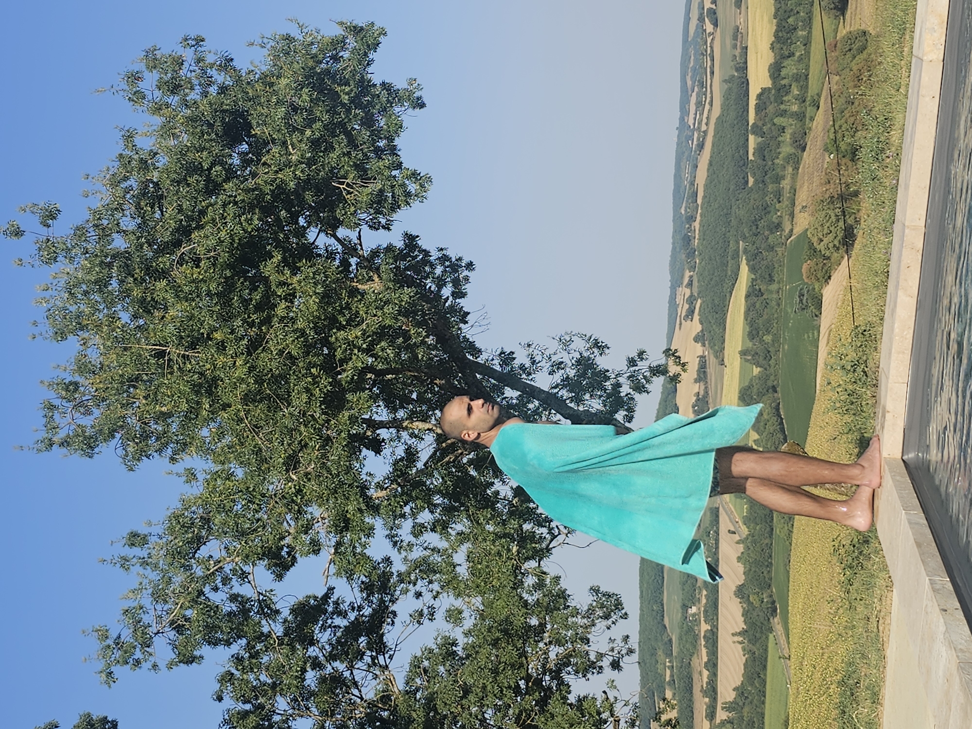 A man is standing on the edge of an infinity pool, draped in a bright blue towel. He is bald and looking to his right. The pool overlooks a rolling landscape of green fields and trees under a clear blue sky. The scene appears to be in Rozès, France, a rural setting with agricultural fields and natural vegetation.
