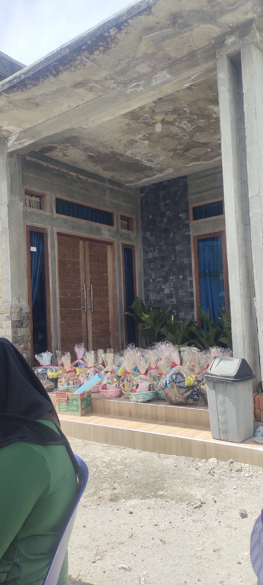 In Simpangantara, Indonesia, on what appears to be a residential property, a scene unfolds before a large wooden double door. Numerous gift baskets, each meticulously wrapped in clear plastic and tied with red ribbon, are arranged on the front steps. These baskets contain an assortment of items, including what appear to be snacks and other goods. To the right of the steps, a large, grey trash receptacle stands, suggesting a functional, everyday setting. Behind the baskets, large green plants add a touch of nature to the entrance. The architecture of the house features exposed concrete beams and columns, hinting at a construction phase or a minimalist design. The doors are flanked by windows with blue curtains, and the exterior wall incorporates a textured dark stone facade. In the foreground, the back of a person wearing a green garment and a dark head covering is visible, seated and facing away from the camera. The ground is a mix of gravel and sand, with some construction materials, like bricks, present near the trash receptacle. The overall impression is a presentation of gifts or offerings at the entrance of a home.