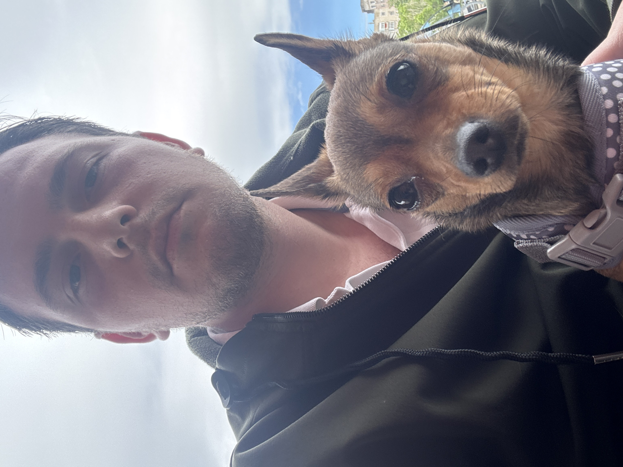 A close-up, angled selfie shows a man holding a small dog. The man has short, dark hair and stubble. He is wearing a dark green jacket over a white shirt. The dog is brown and black with a grey and white polka-dotted harness. The background is a cloudy sky and a blurred building, potentially located in the city of Plovdiv, Bulgaria.