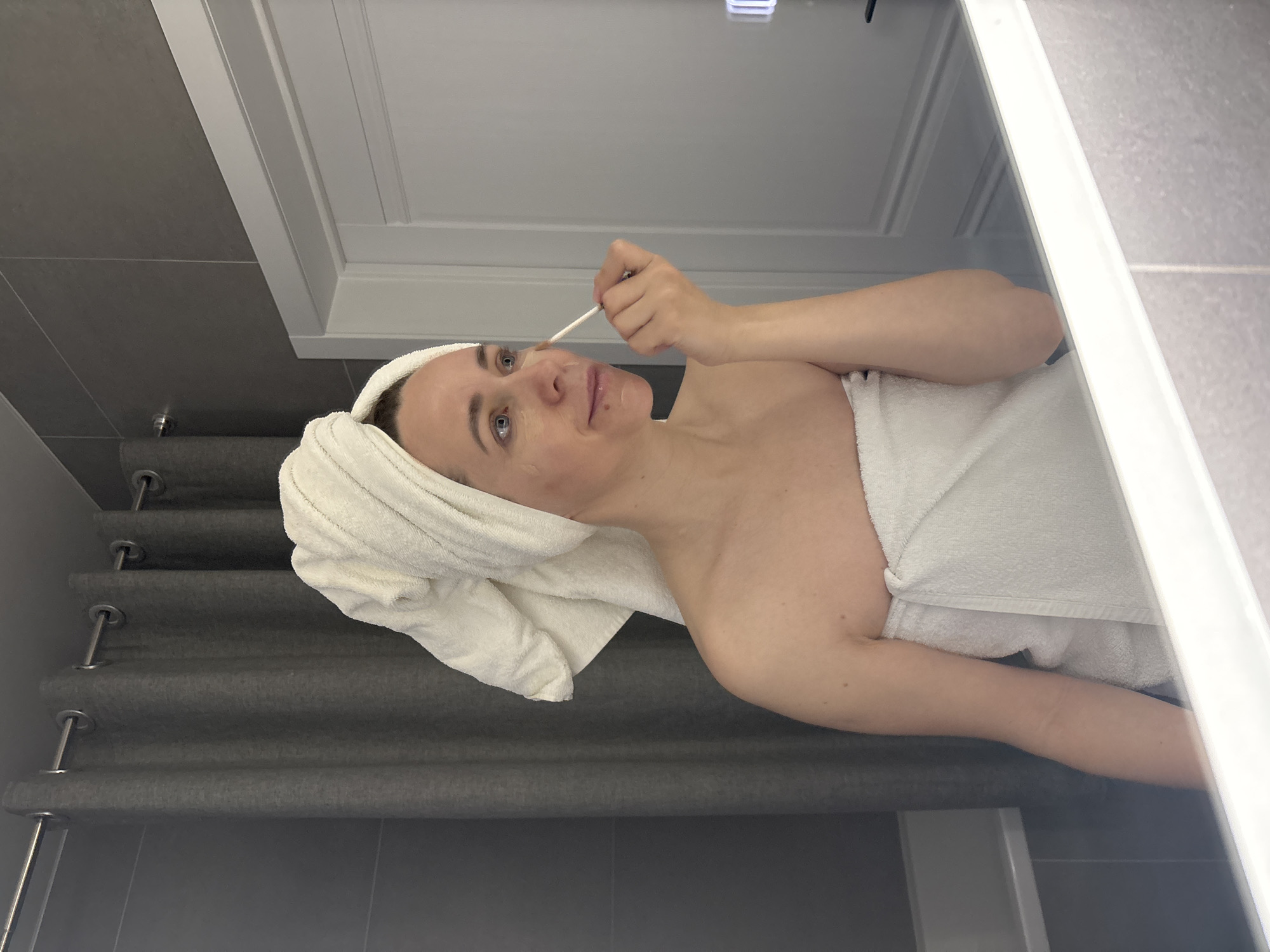 A woman, appearing to be in her 30s or 40s with light skin and blue eyes, is depicted engaged in a skincare routine within what appears to be a modern bathroom. Her light brown hair is neatly wrapped in a white terrycloth towel, and she is also draped in a matching white towel around her body. The image is rotated 90 degrees clockwise, making her appear to be lying on her back, but she is likely standing or sitting upright, facing a mirror.
She is focused on applying a substance to her face, specifically below her left eye, using a small, white-tipped applicator stick, possibly a cotton swab or a cosmetic tool, held delicately between her right thumb and forefinger. Her facial expression is neutral and concentrated. There are small, subtle marks or blemishes visible on her face, which could be part of the treatment process.
The background reveals typical bathroom elements. Grey tiled walls and flooring are prominent, suggesting a clean, minimalist aesthetic. To her left, grey fabric blinds with horizontal pleats and visible metallic grommets are seen, potentially covering a window. A white architectural feature, possibly a door frame, mirror frame, or part of a vanity, frames the upper and right edges of the scene. A square, white light fixture is reflected in the upper background, indicating artificial indoor lighting.
The overall setting suggests a private, self-care moment. The image provides no visible text. This scene takes place in Chaikovsky, Russia.
