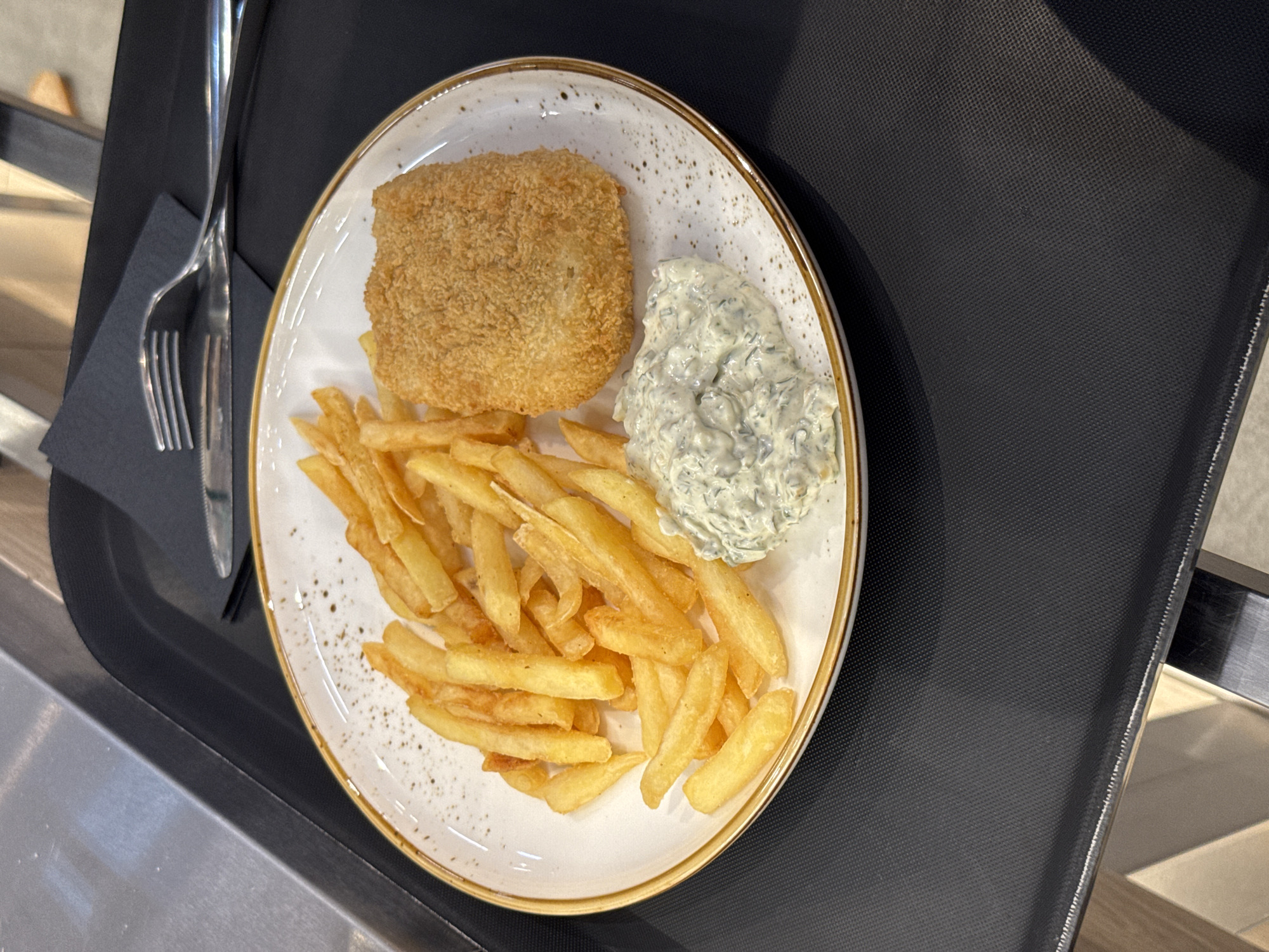A plate of fish and chips, with a portion of tartar sauce, is presented on a black tray. The plate has a speckled pattern and a gold rim. The fish appears to be breaded and golden brown, while the french fries are thick-cut and also golden. A dark grey napkin is under a fork and knife. The setting is an indoor environment, possibly a restaurant, with a metal countertop and wooden elements visible in the background. The image does not provide any specific information to locate it in Lakhdenpokhya, Russia.