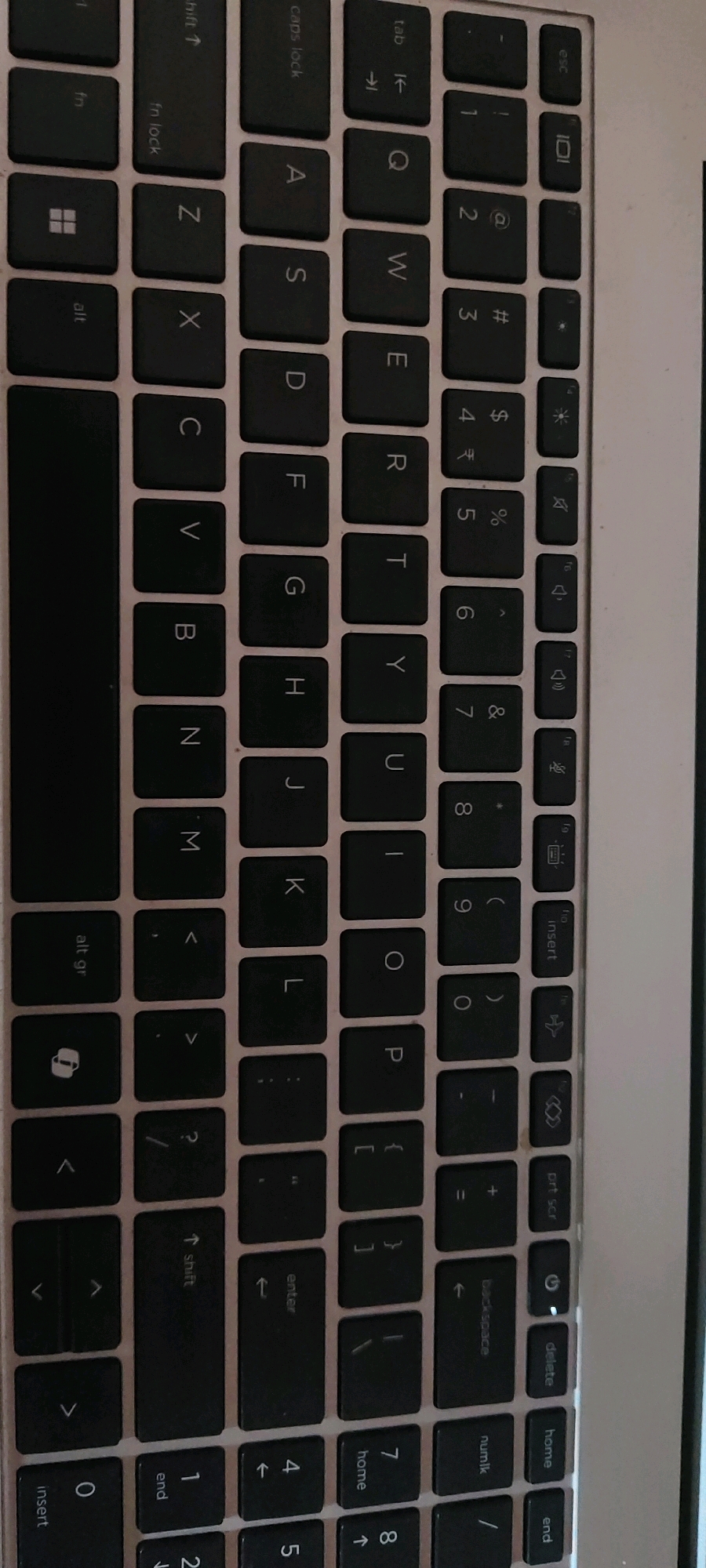 The image shows a close-up view of a laptop keyboard. The keyboard is black with silver lettering on the keys. The keys are arranged in a standard QWERTY layout, with additional function keys and symbols. The orientation of the image is such that the keyboard is vertical, and the perspective is slightly angled, revealing the side of the laptop.
There are no people, animals, or specific environmental features visible in the image, other than the keyboard itself and the metallic casing of the laptop. The lighting appears to be artificial, casting shadows that highlight the texture of the keys. The overall impression is of a clean, functional electronic device. The request for location in Kuntanahosalli, India, cannot be fulfilled as the image provides no geographical context.