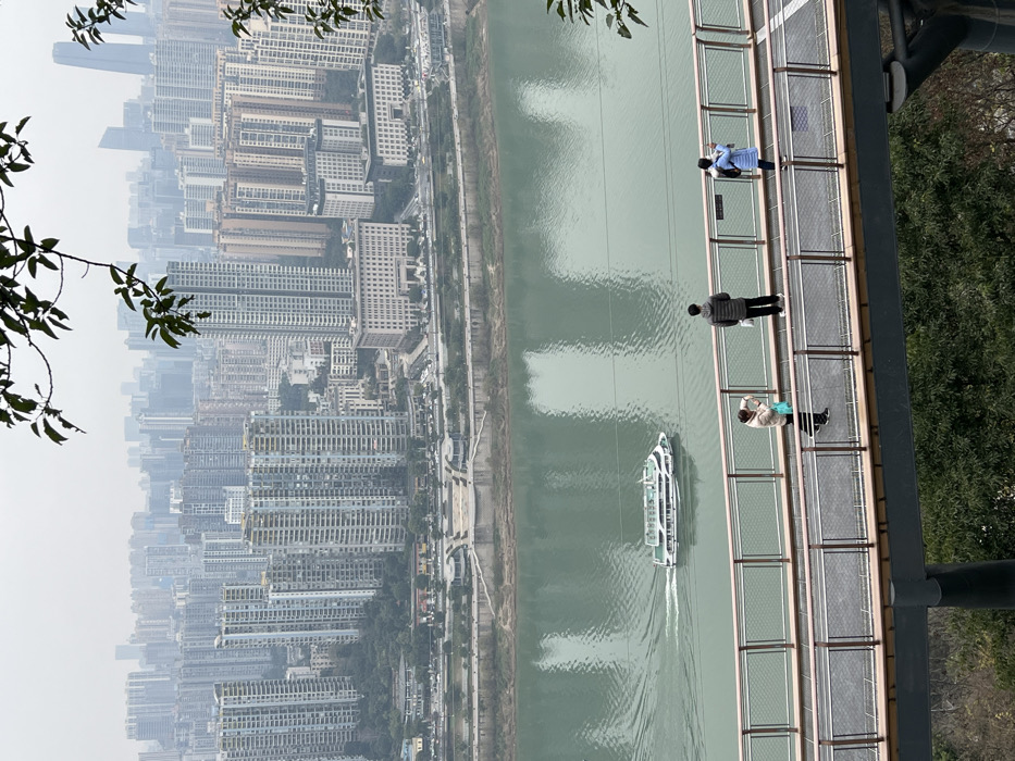 An elevated view showcases a river and cityscape. The river, navigated by a sightseeing boat, is bordered by a dense urban area filled with tall residential buildings. A pathway elevated above the river is occupied by three pedestrians. A tree branch with green leaves is visible in the upper left corner. The opposite side is vegetated, as well.