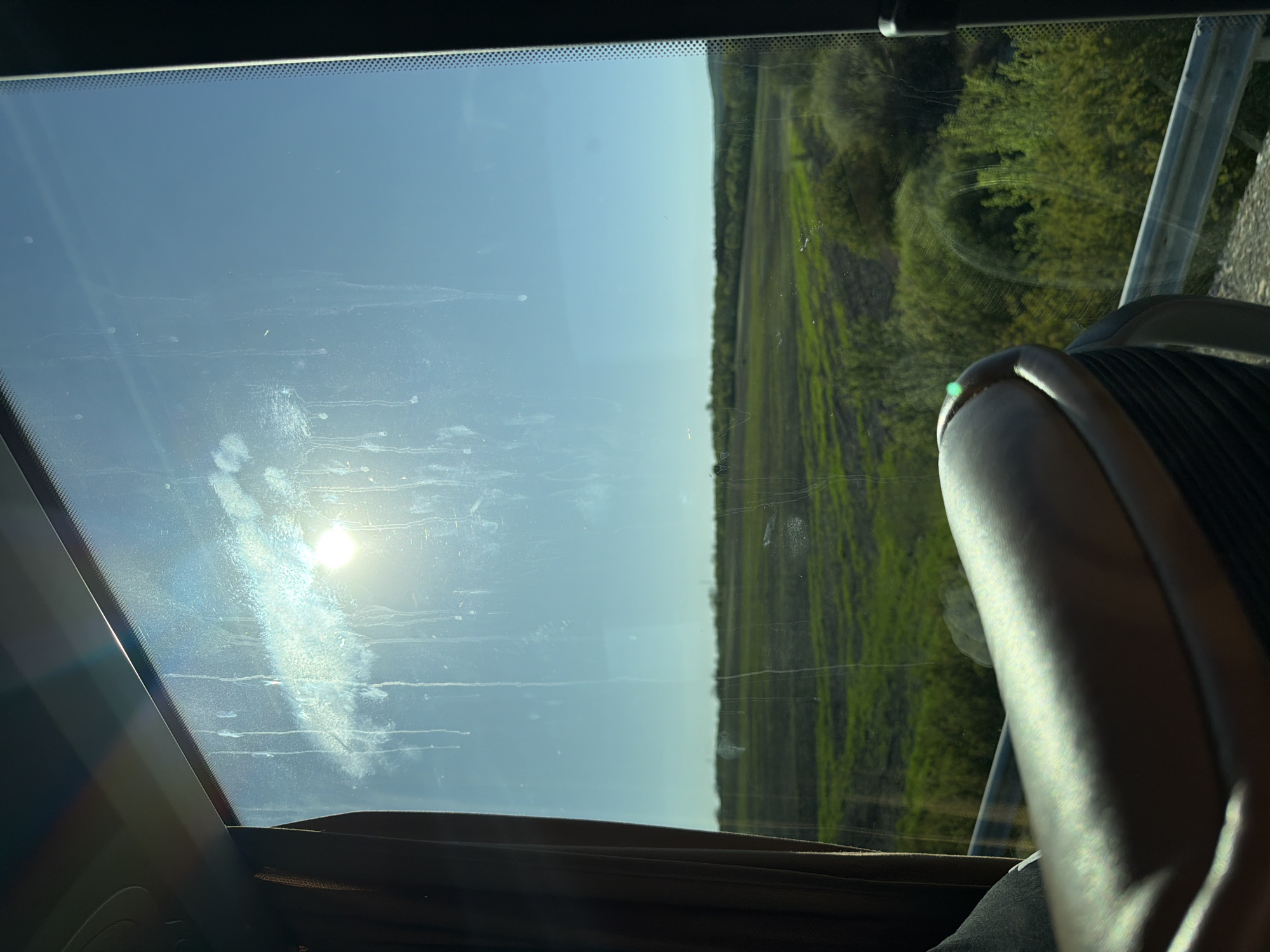 The image depicts a view through a dirty window, likely from inside a bus or vehicle. Outside, there is a field or landscape with green vegetation. A portion of a seat is visible in the foreground. The location, Malko Tranovo, Bulgaria, cannot be confirmed from the visual data alone. No people are visible. The sun is shining through the window creating glare.