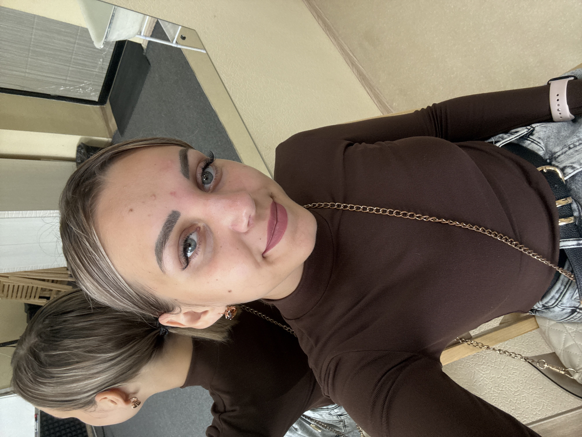 A young woman with blonde hair and blue eyes is pictured in an indoor setting. She is wearing a brown long-sleeved turtleneck shirt and light-colored jeans. She has a gold chain bag strap across her chest. Her reflection is visible in a mirror behind her, which also shows a doorway with a mat on the floor and a piece of furniture that appears to be a shelf unit. The walls are a light yellow color. The scene appears to be a personal space, possibly a dressing room or a home. There is no indication of the specific location within the city of Yuzhno-Sakhalinsk, Russia.