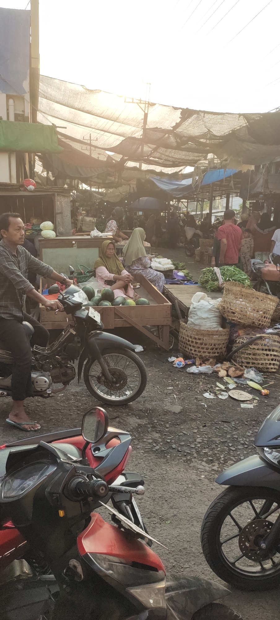 In Karangtowo, Indonesia, a bustling market scene unfolds under a hazy sky. A man, wearing a plaid shirt and dark pants, rides a black motorcycle from left to right in the foreground. He is heading towards a stall where two women, one in a green hijab and pink clothing and the other in a light green hijab and patterned dress, are seated. The stall displays a large quantity of watermelons. Behind them, other vendors and shoppers are visible amidst various market stalls covered with tarps. Various goods are being sold, including what appear to be vegetables and woven baskets filled with produce. The ground is a mix of dirt and asphalt, littered with some trash. In the foreground, parts of two other motorcycles are visible, one red and black and another gray. The lighting suggests it is either early morning or late afternoon.