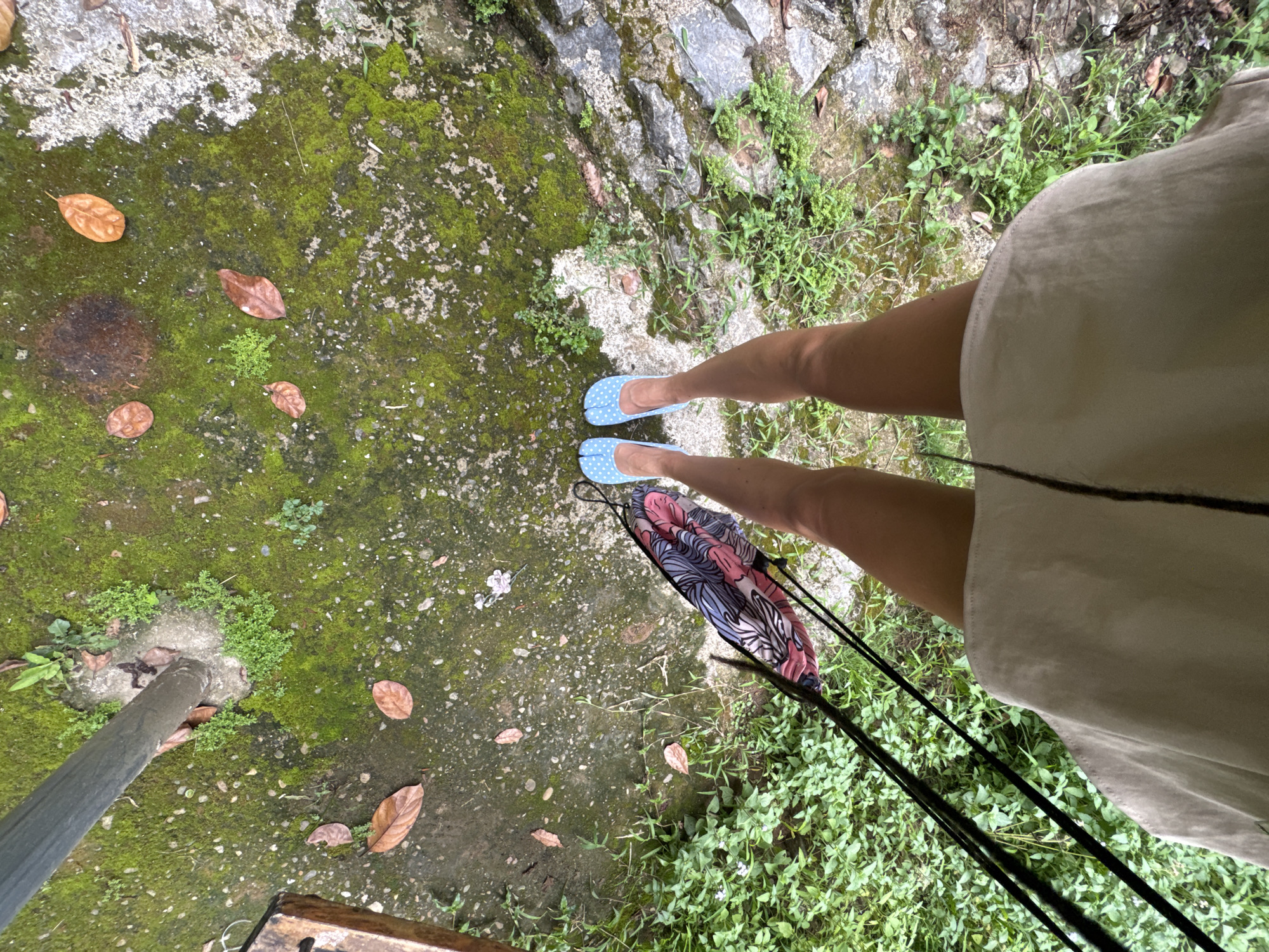 This high-angle shot captures a pair of legs from the knees down, positioned on moss-covered ground with scattered dry leaves and sparse vegetation. The individual is wearing light blue, polka-dotted split-toe shoes. A patterned bag with pink, blue, and white designs is held by black straps, hanging beside the right leg. The lower portion of a beige garment, possibly a dress or skirt, is visible in the foreground. The surroundings suggest an outdoor, possibly natural or unkempt urban, environment. There is no specific indication of Cẩm Thạch Commune, Vietnam, within the image itself.