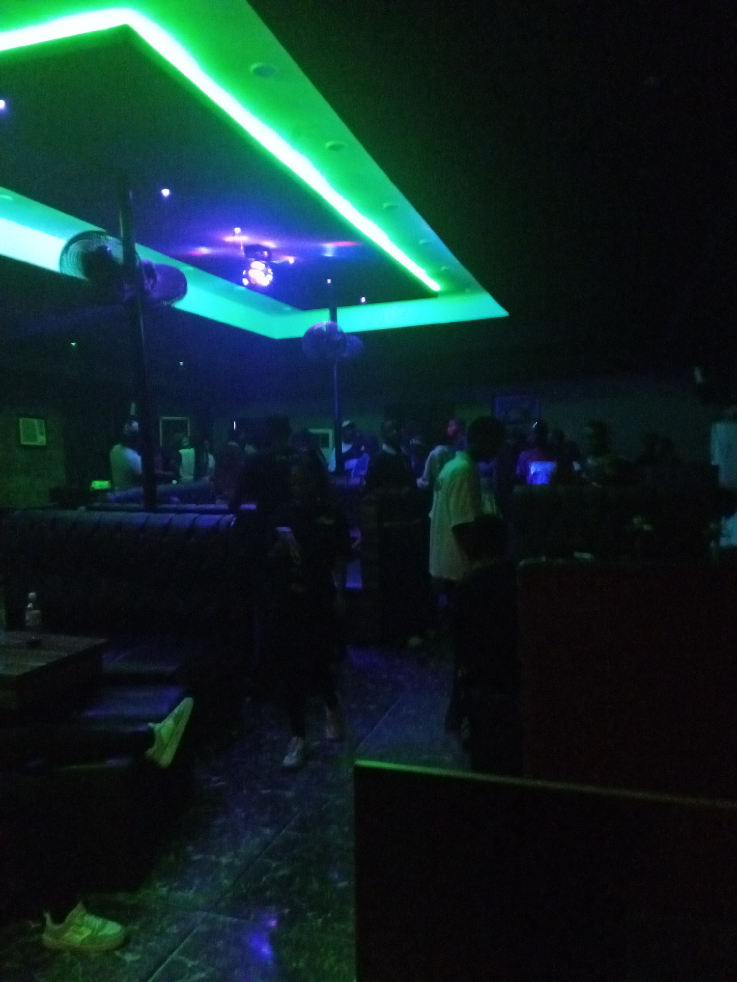 The image shows the interior of what appears to be a nightclub in Lokoja, Nigeria. The lighting is predominantly dark, with a green fluorescent light outlining a section of the ceiling. A disco ball is visible in the center of the ceiling. There are several people in the room, some standing and others seated on a dark leather sofa. Details of individual faces are obscured due to the low light conditions. Shoes are on the floor. Additional objects such as tables and what appears to be an air conditioner are discernible.