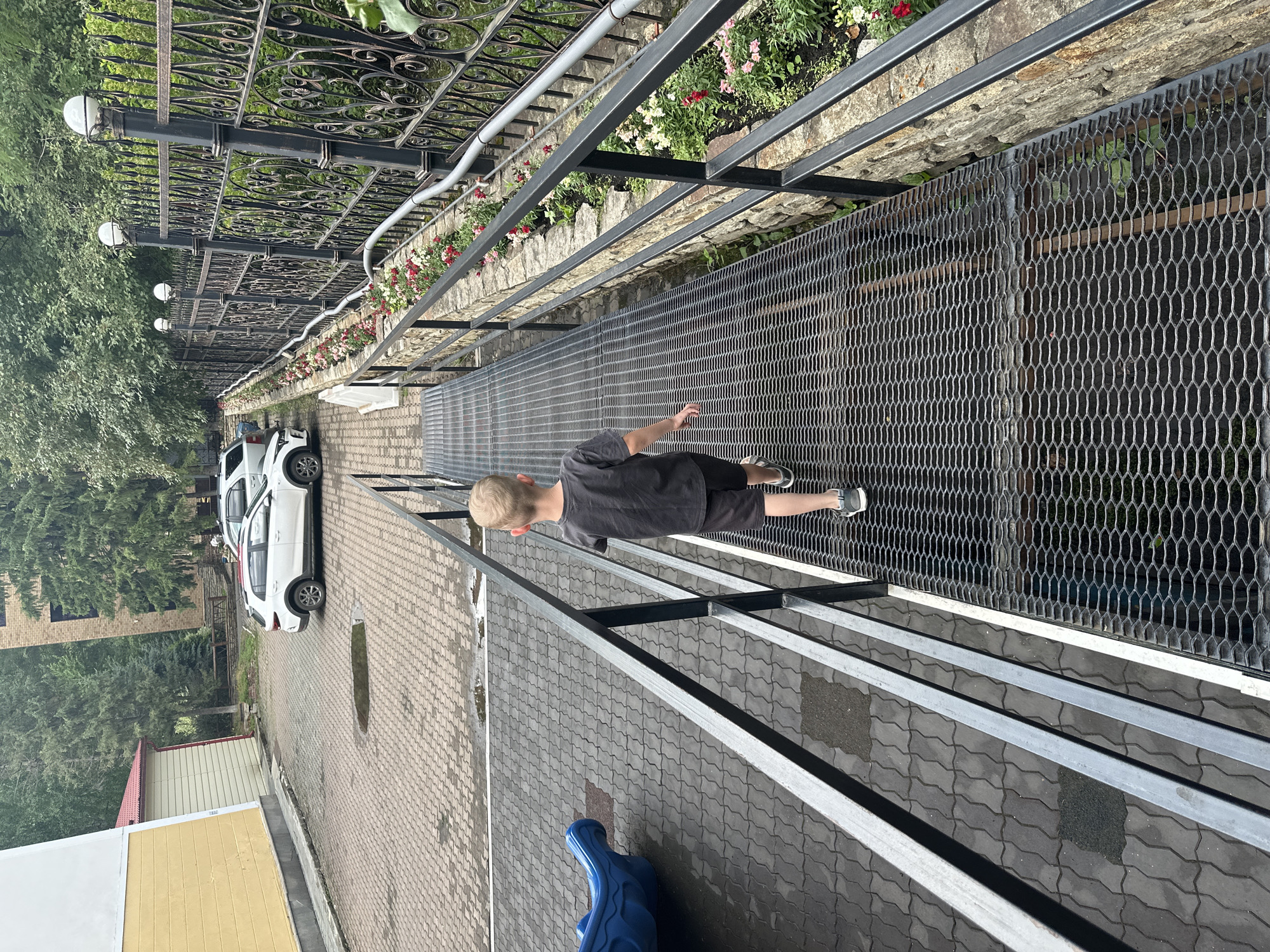 A blond-haired boy wearing a dark t-shirt and shorts walks away from the camera on a metal grate ramp. The ramp has a metal railing on both sides. To the boy's left, there is a blue playground slide and a paved area with interlocking bricks and some puddles. In the background, there are two white cars parked in a lot, a yellow building with a red roof, a brick building, trees, and an ornate black metal fence with glass globe lights. The fence is lined with a flower bed containing red and white flowers. The scene appears to be an outdoor area near buildings and a playground, possibly in the city of Belokurikha, Russia, given the architectural style and landscaping.