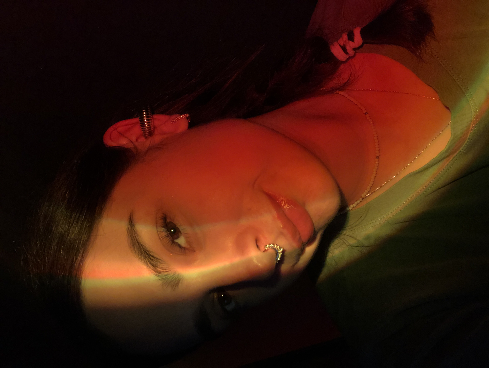 In the city of Noviy, Russia, a young person is depicted in a close-up, dimly lit portrait. A strong beam of light, appearing to be from a colored projector or prism, casts a vertical stripe of green, yellow, and orange light across their face, starting from their forehead and extending down to their shoulder. The person has dark hair, styled in a braid that is partially visible on the right side of the frame. They are wearing a light green t-shirt and multiple necklaces, one of which appears to be made of small beads, and another a delicate gold chain. The person has a septum piercing with a detailed, silver-colored ring, and a ribbed ear cuff on their right ear. The background is predominantly dark, with the exception of the colored light.
