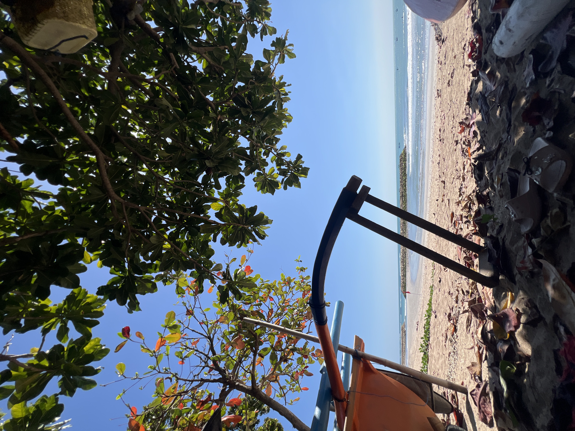 A low-angle, natural light, outdoor shot captures a scene on a sandy beach in Kuta, Indonesia. The foreground is dominated by fallen leaves scattered across the sand, partially obscuring some scattered debris including what appear to be shoes. To the left, the orange and blue structures of a boat are visible, propped up by wooden poles. Beyond the boat, the sand leads to the ocean, where gentle waves are breaking on the shore. Stone jetties extend into the water, and a small red flag is planted on the beach. Several small boats are visible on the water. The sky above is clear and blue, with the branches and leaves of two trees providing a canopy over the scene. One tree has vibrant green leaves, while the other displays a mix of green, yellow, and red leaves.