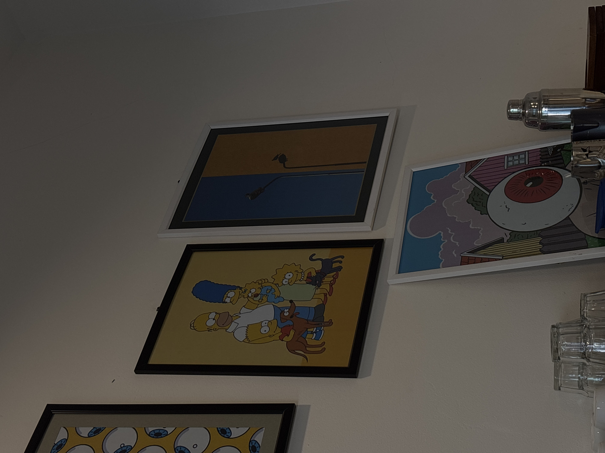 The image displays a collection of framed artwork hung on a plain white wall. On the far left, a framed artwork features a repeating pattern of stylized eyeballs with blue irises. To its right, a larger framed picture depicts the Simpson family, including Homer, Marge, Bart, Lisa, Maggie, and their pets Santa's Little Helper and Snowball II, against a yellow background. Next to the Simpson artwork is a framed piece with a split background, one half deep blue and the other a warm ochre, featuring silhouetted streetlights with birds perched on them. Below these, a framed artwork with a white border showcases a drawing of a pink house with a yellow roof, a stylized eye on a stalk, and a cloudy sky. In the lower right corner of the image, a stainless steel cocktail shaker and several stacked glasses are visible on a shelf. The scene appears to be an indoor setting, possibly a cafe or a home, given the display of art and drinkware. There is no indication of this scene being located in Semarang, Indonesia.