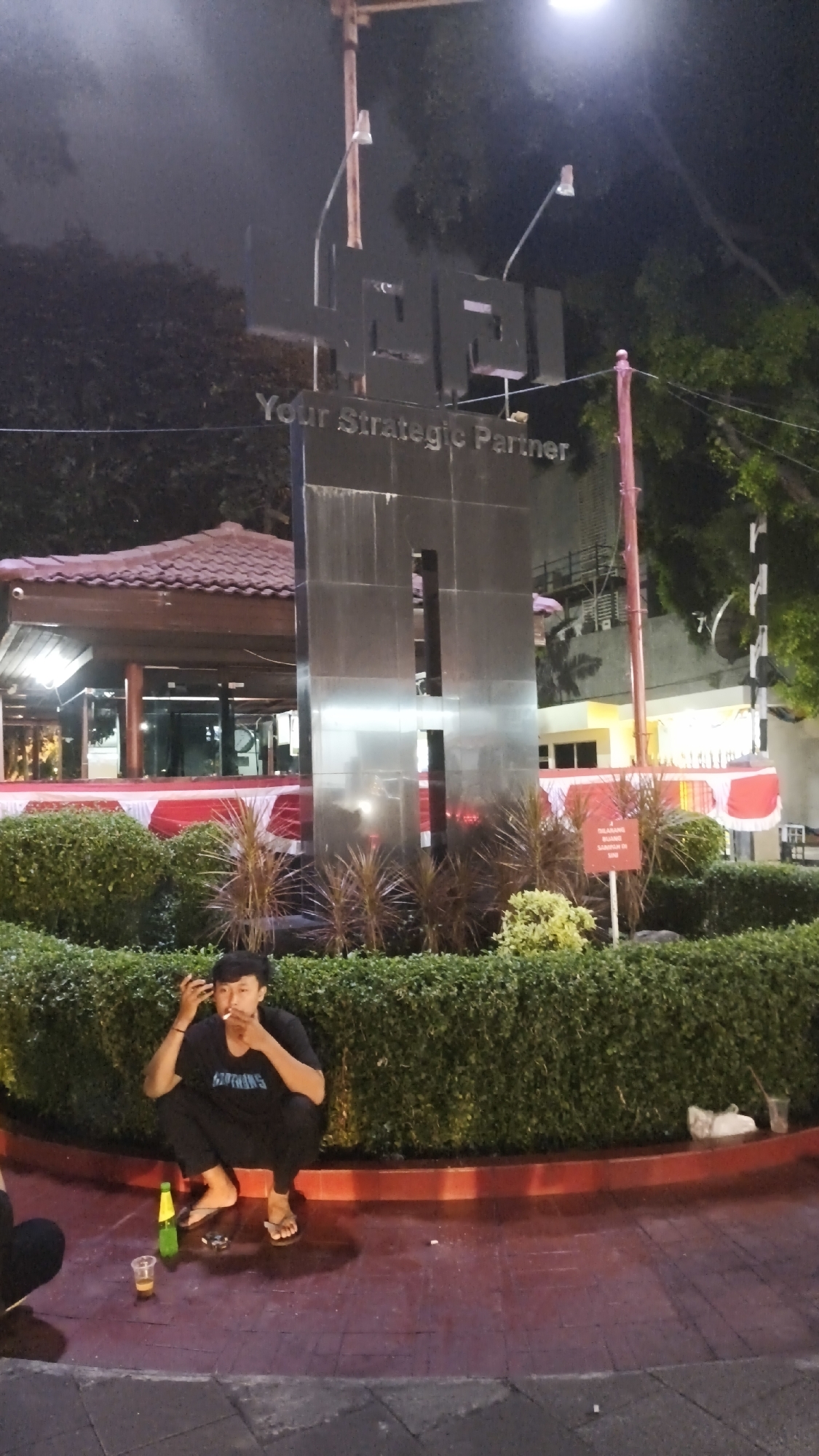 An outdoor nighttime scene in South Jakarta, Indonesia, features a man squatting on a red tiled surface in front of a neatly trimmed hedge. The man, wearing a black t-shirt and pants, is smoking a cigarette and appears to be holding his hand to his face. To his left, there is a green bottle and a plastic cup containing a beverage. Behind the hedge, a large, dark, rectangular monument with cutouts stands prominently. The monument has the text "Your Strategic Partner" inscribed on it. In the background, a building with a red tiled roof and red and white draped decorations is visible. Trees are present in the upper portion of the image, and streetlights illuminate the area. The overall lighting suggests it is evening or night.