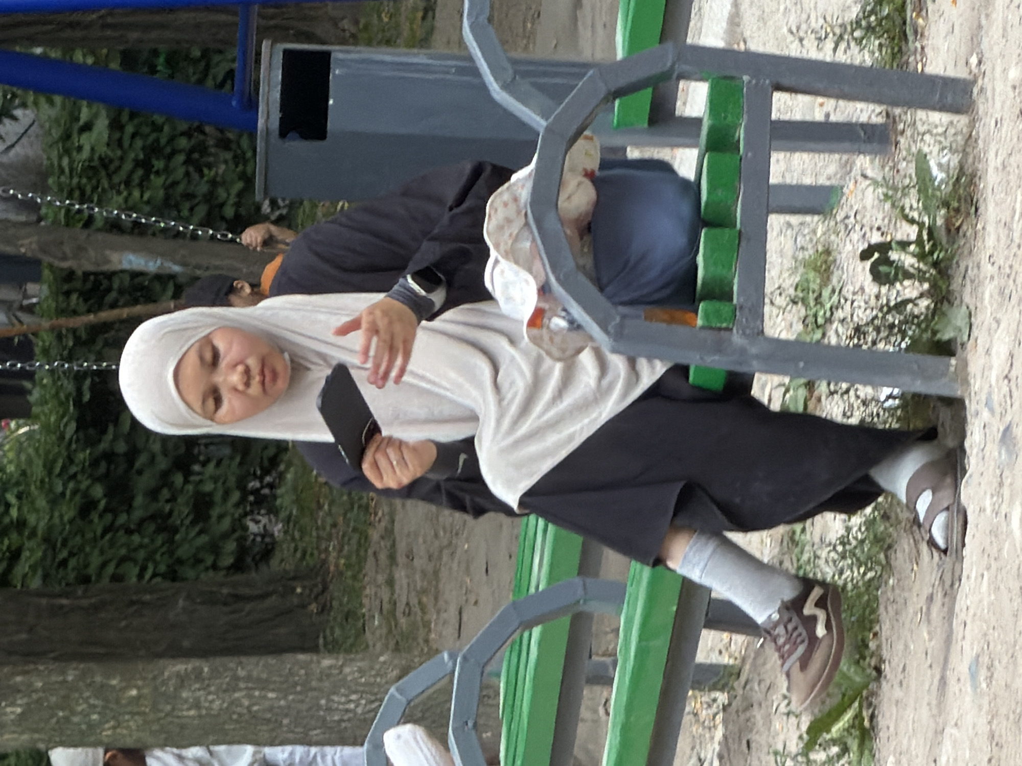 In an outdoor urban setting, a woman wearing a light-colored hijab and a dark abaya sits on a green park bench. She is holding a dark rectangular object, possibly a phone or wallet, in her right hand, which is extended as if she is about to gesture. Her left hand is near the object. She is wearing a smartwatch on her left wrist and appears to be looking down. Her legs are crossed, with her right foot wearing a brown athletic shoe and grey sock, and her left foot wearing a brown sandal and white sock. Another person, partially visible and wearing a white garment and hijab, is standing to the left behind a tree. In the background, there is a swing set with chains and a blue metal frame, a grey trash can, and trees with green foliage. The ground is covered in dirt and scattered leaves.