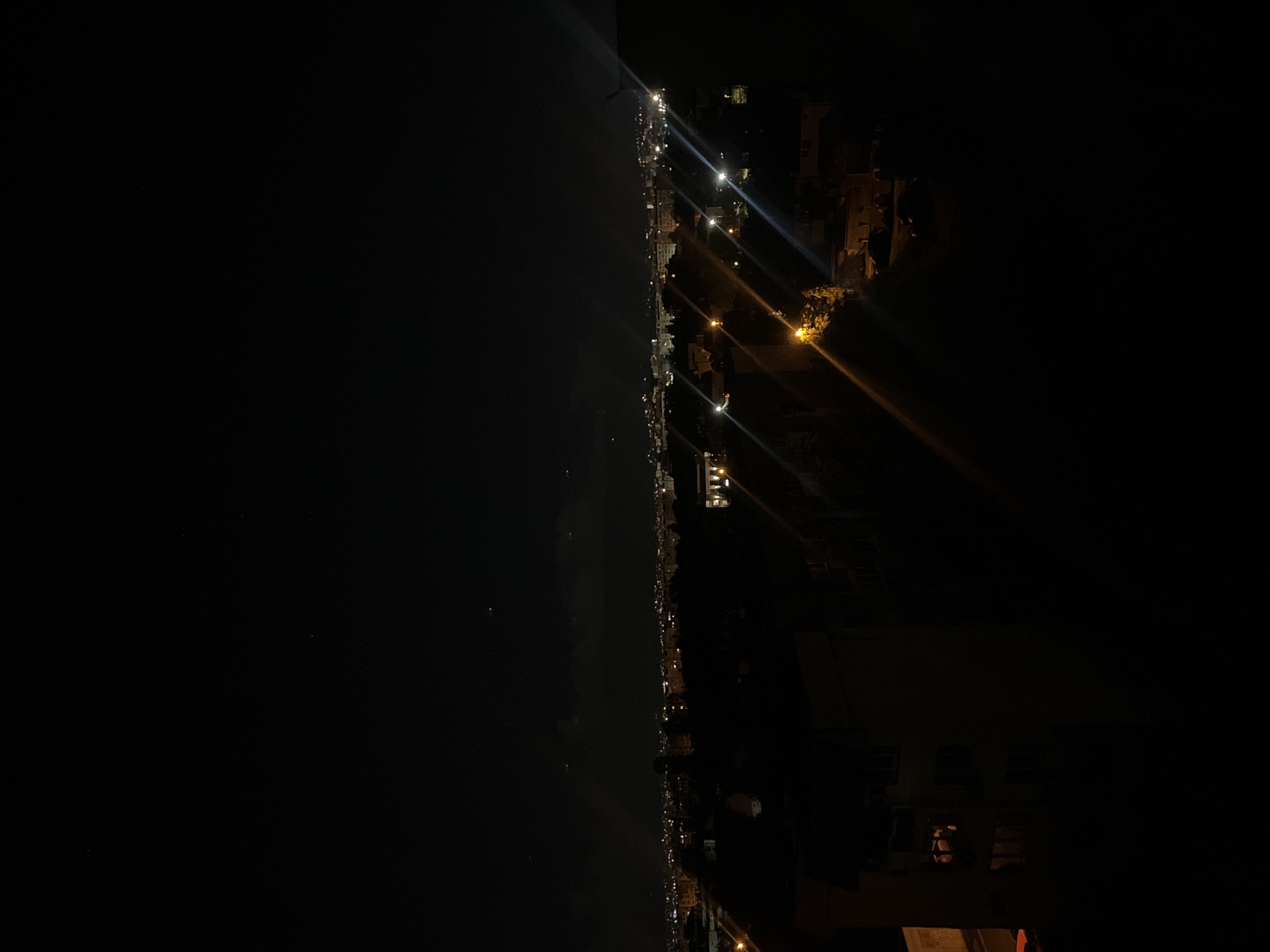 The image depicts a nighttime cityscape. A dark body of water stretches along the left side. A line of lights indicates a road or pathway that traces along the edge of the water. Buildings stand on the right side of the image, illuminated by streetlights and interior lights. The environment is dark, with the illumination provided by artificial light sources. There are no discernible people in the image. The image is likely taken from a high vantage point, possibly a building or hill overlooking the cityscape. There is no information about Sofia, Bulgaria.