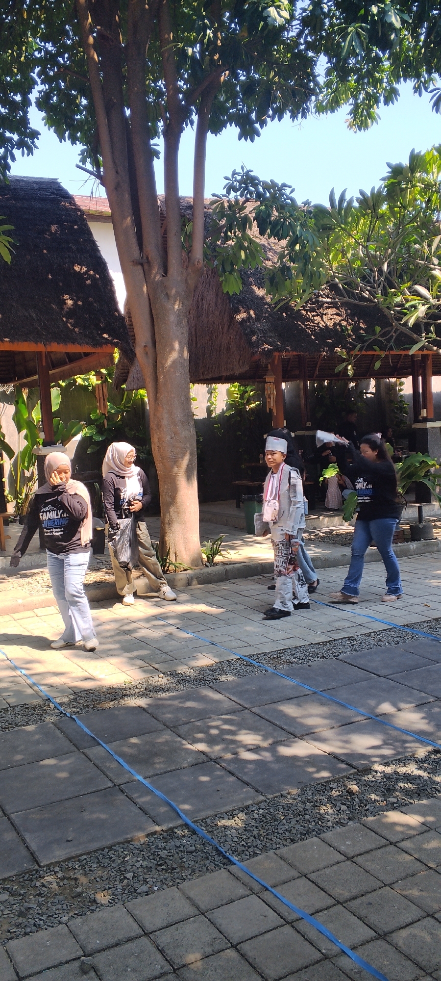 In Talise, Indonesia, under a clear blue sky and dappled sunlight, a group of people are gathered outdoors. A large tree with a rough, textured trunk stands prominently in the foreground, its branches extending upwards and outwards, creating shade. Behind the tree, thatched-roof structures, likely gazebos or outdoor seating areas, are visible, suggesting a recreational or dining space.

Several individuals are present, walking and standing. On the left, two women are walking. The woman in the foreground wears a light-colored hijab and a dark t-shirt with the word "FAMILY" in white lettering, paired with light-washed jeans and white shoes. To her left, another woman in a light hijab and dark long-sleeved shirt walks, holding a black bag and a white object.

To the right of the tree, a young person in a light-colored outfit, possibly a uniform with a white head covering and a sash, stands and appears to be looking to their left. Further to the right, another woman in a black t-shirt with "FAMILY" visible again, and blue jeans, is walking away from the camera.

The ground is paved with square stone tiles, interspersed with strips of gravel, with thin blue plastic strapping laid across the paving in some areas. The overall scene suggests a casual outdoor gathering or event in a warm climate.