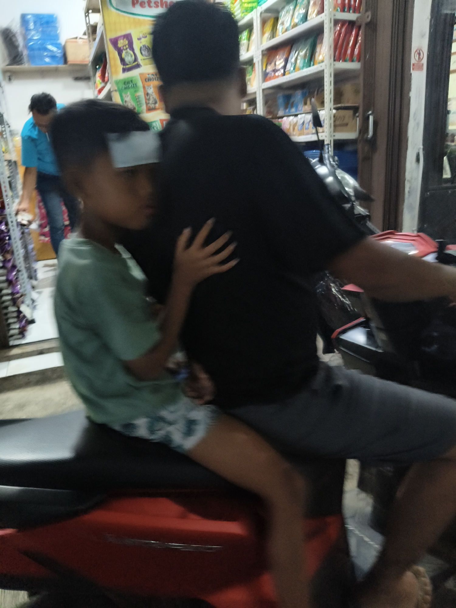 A medium shot shows two individuals, likely a child and an adult, riding on a red and black scooter in what appears to be a street market or shop in South Tangerang, Indonesia. The child, in the foreground and on the left, wears a light green t-shirt and patterned shorts. The adult, on the right and behind the child, is dressed in a black t-shirt and gray pants. The child is looking towards the right, while the adult's head is turned away from the camera. Shelves stocked with various items, possibly pet food given the "Petshop" sign visible on a yellow banner, are in the background. A blue plastic container is also visible on a shelf to the left. The scene is dimly lit, suggesting it might be evening or indoors.