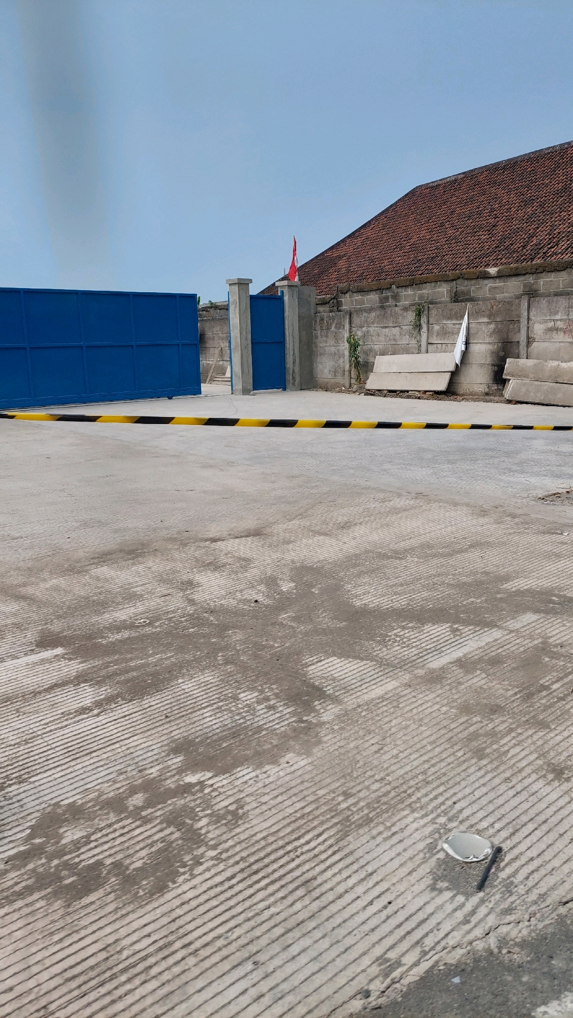 The image depicts an outdoor scene with a bright sky. In the foreground, a textured concrete surface is visible, with diagonal grooves and some water stains. A yellow and black striped barrier lies horizontally across the scene. Behind the barrier, a large blue gate stands to the left. To the right of the gate are two concrete pillars, one of which has a small red and white flag attached to its top. A long, low building with a tiled roof occupies the background, with a grey concrete wall running along its base. Several concrete slabs are stacked against the wall. The overall impression is one of an industrial or commercial area, possibly an entrance to a facility, located in Sidoarjo, Indonesia.