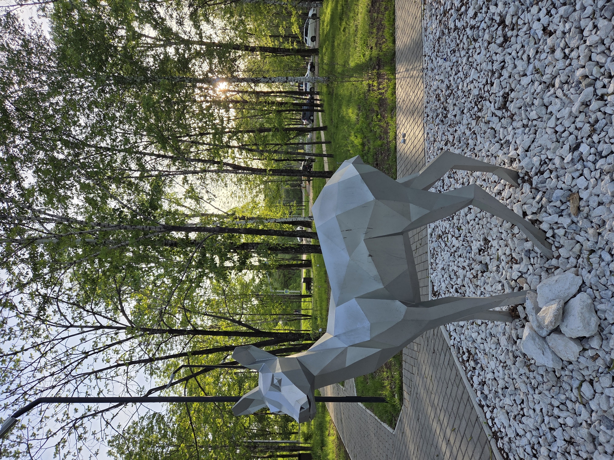 The image depicts a low-poly art sculpture of a deer in a park setting. The sculpture is made of a light-colored material, possibly metal or concrete, and has a geometric, faceted appearance. It stands on a bed of small white rocks next to a paved walkway. The background features a row of trees with green leaves, through which sunlight is filtering. Several vehicles are parked along the street behind the trees. The location is Birobidzhan, Russia, based on available information. No people are visible in the image. The overall environment suggests a public park or recreational area.