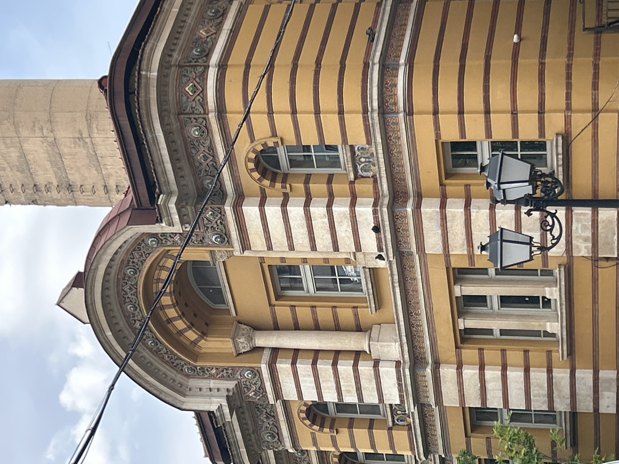 The image shows the exterior of a building in Sofia, Bulgaria. The architectural style features elaborate ornamentation, including stripes, columns, arched windows, and decorative patterns. A tall, cylindrical structure stands adjacent to the building, contrasting with the building's ornate facade. A streetlight is mounted on the building's facade. The sky is visible in the lower-left corner.