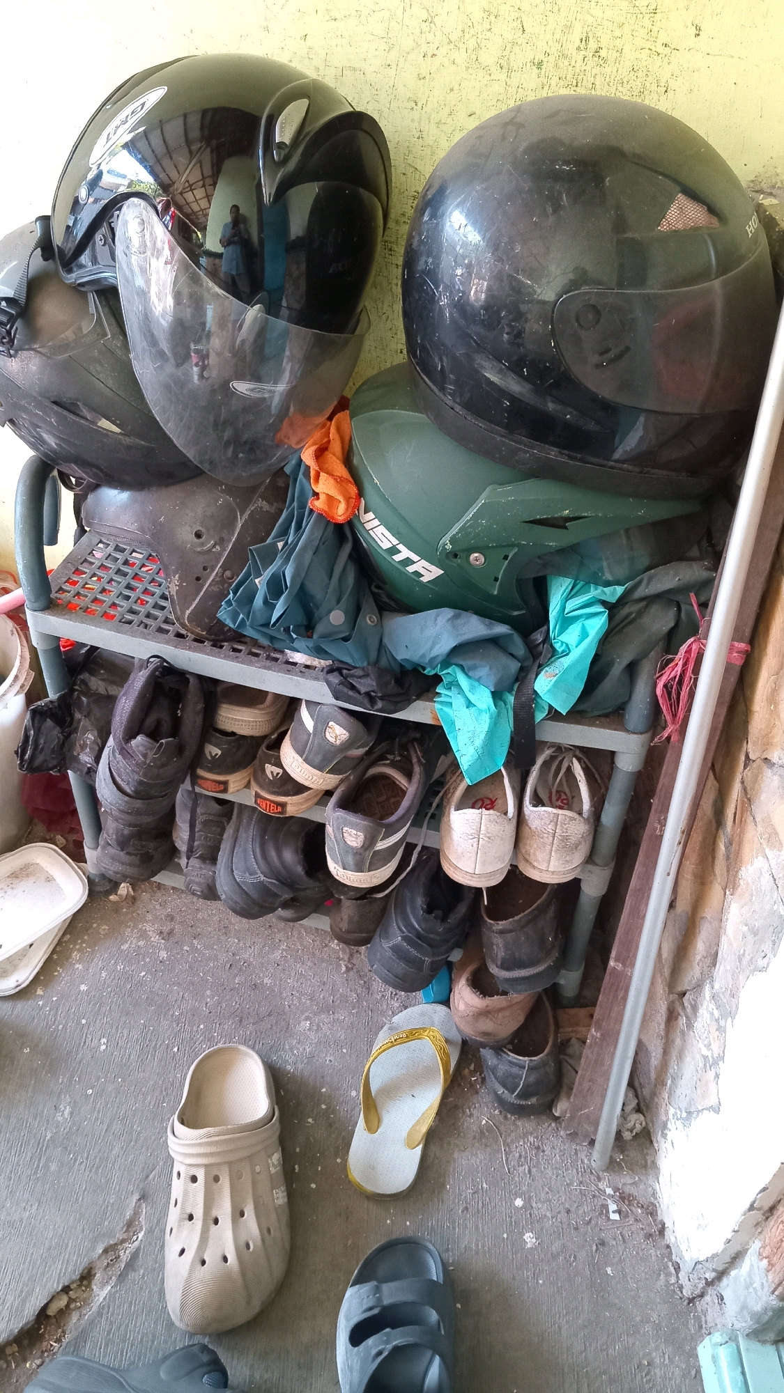 The image depicts an outdoor scene in Randuagung, Indonesia, likely near a residential area or workshop, given the presence of motorcycles and storage. In the foreground, several pairs of footwear are scattered on a concrete floor, including a beige clogs-style sandal, a yellow and white flip-flop, and a dark gray sandal. Behind these, a multi-tiered gray plastic shoe rack is filled with various shoes and boots. On the top shelves of the rack, a collection of motorcycle helmets is stacked. These include a black full-face helmet with a reflective visor, another black helmet with a more rounded design, and a green motocross-style helmet. Several pieces of clothing, including what appear to be dark blue and orange fabric items, are draped over the helmets and the rack. The background shows a textured wall, possibly brick or plaster, and a wooden plank leaning against it. The overall impression is one of storage for outdoor gear.