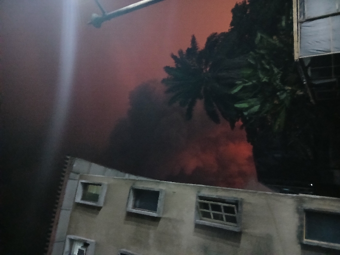 An apartment building with multiple windows stands against a backdrop of reddish smoke, possibly from a fire. A palm tree is partially visible to the right of the building. An overhead light fixture extends into the frame from the top-left corner. The scene appears to be taken at night or in low-light conditions.